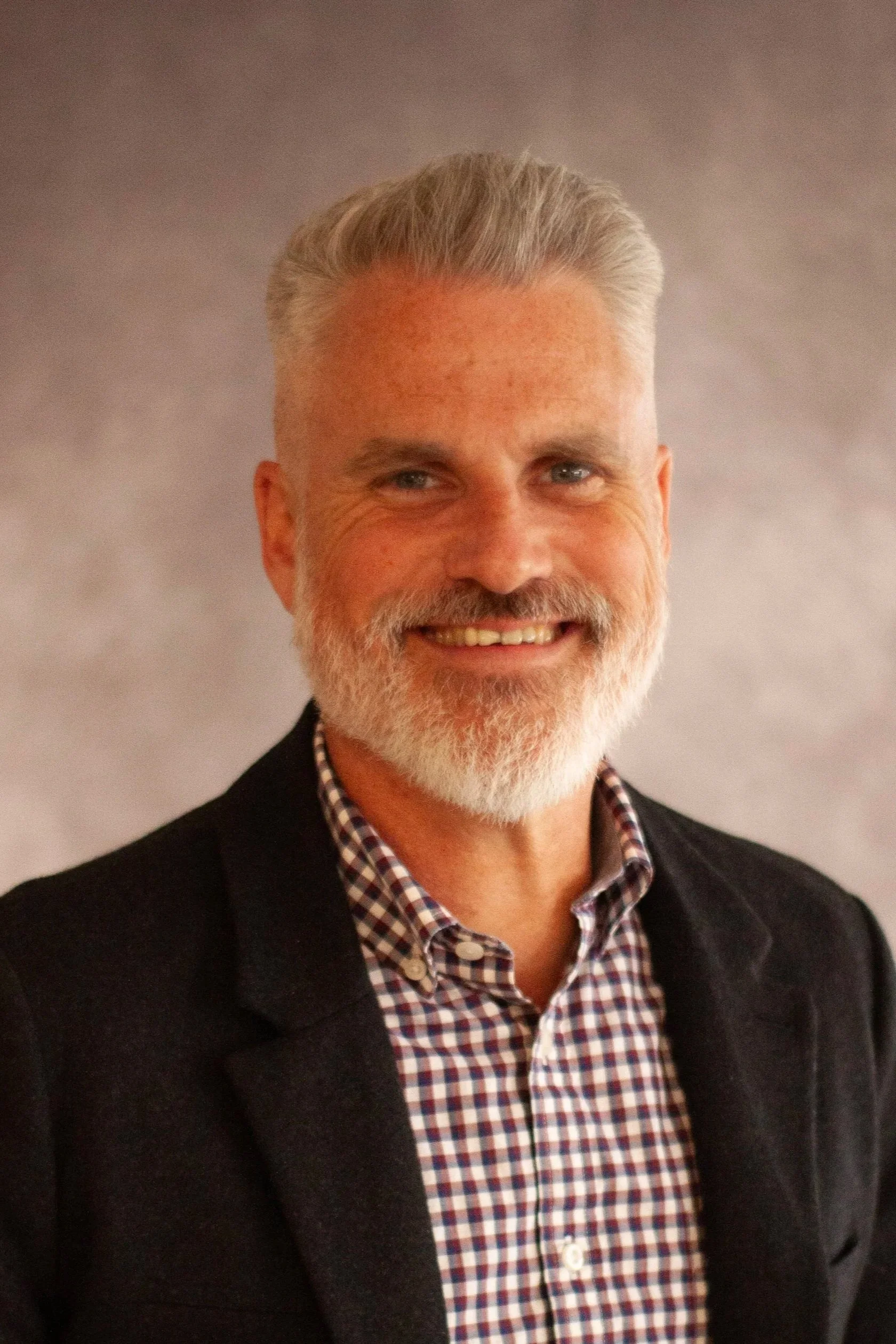 A middle-aged man with gray hair and a beard smiling, wearing a dark blazer over a checkered shirt, standing against a neutral background.