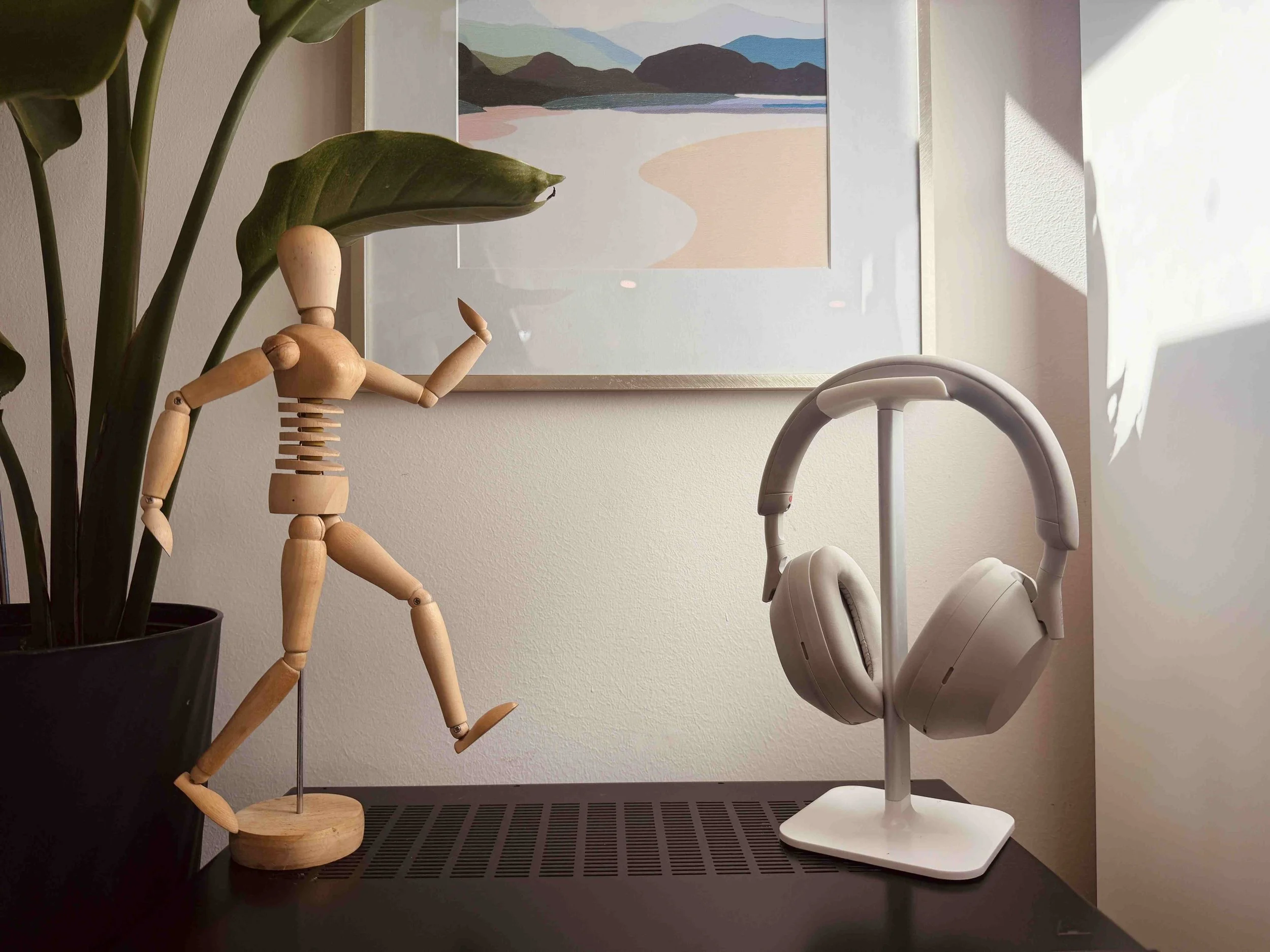 A wooden artist mannequin waving while standing on a flat surface next to a potted plant with large green leaves, and a pair of beige over-ear headphones resting on a headphone stand. There is a framed abstract art piece on the wall in the background, and sunlight is shining into the room creating shadow patterns on the wall.