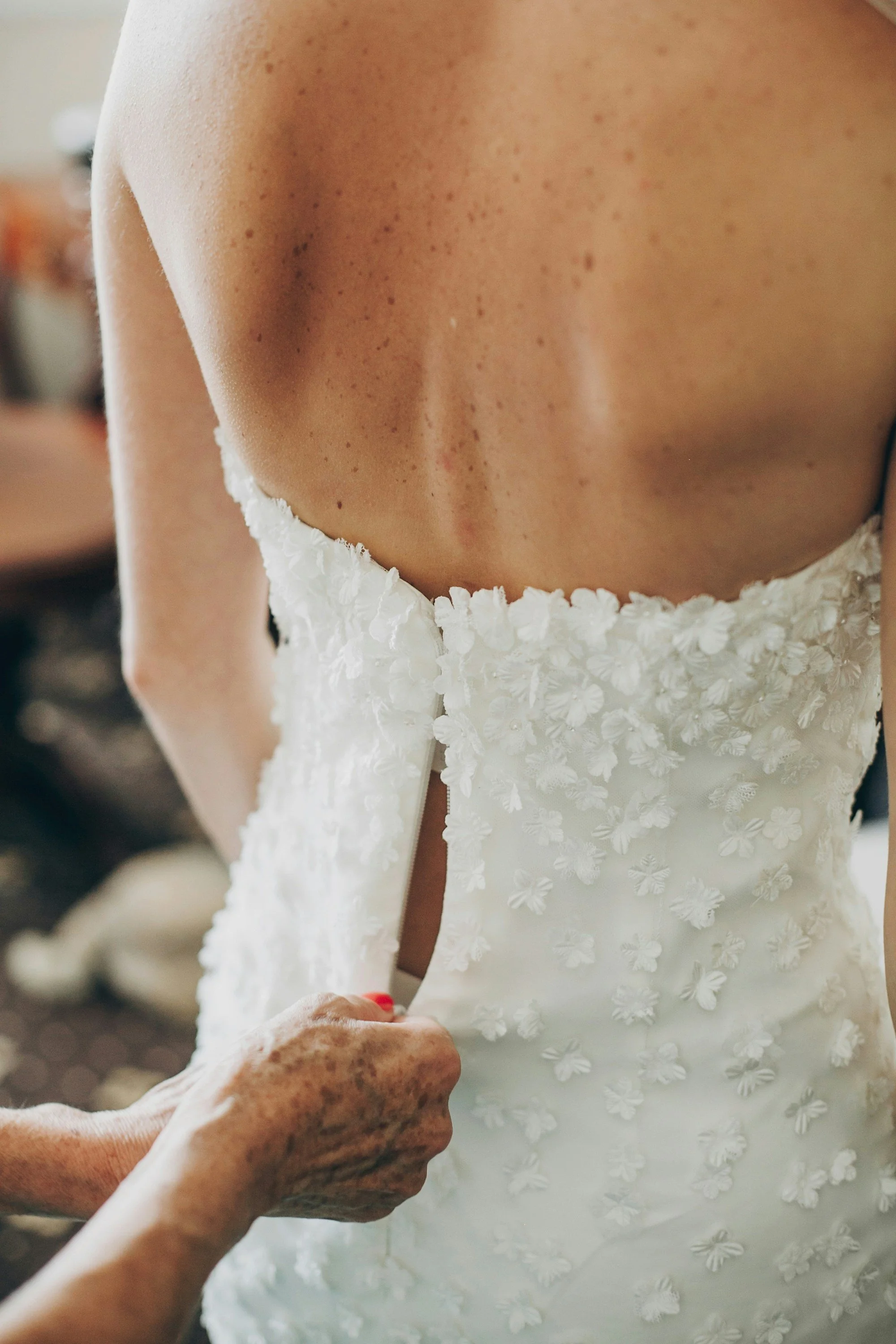 A bride in a white wedding dress with floral applique is being buttoned up at the back, showing her upper back and shoulders.