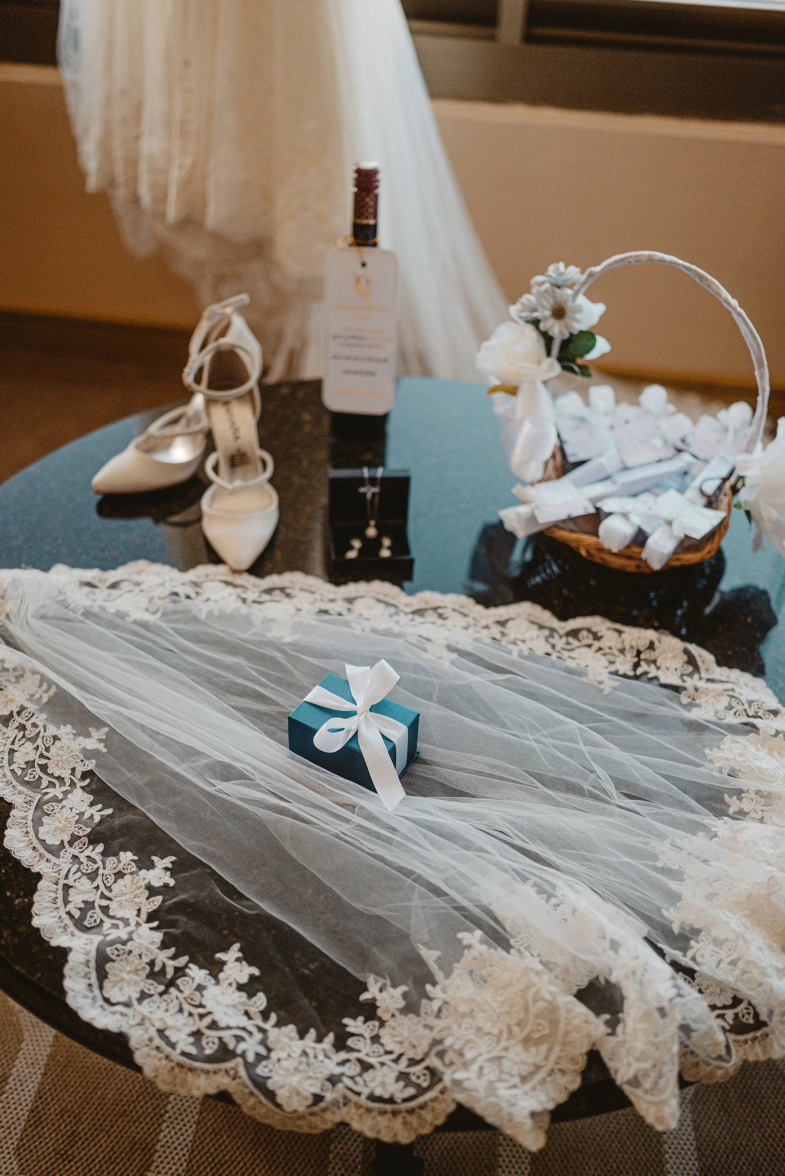 Wedding accessories on a black table, including a white veil with lace, a small blue gift box with a white ribbon, white bridal shoes, a necklace and earrings display, a bottle of wine with a tag, and a basket with white chocolates and flowers.