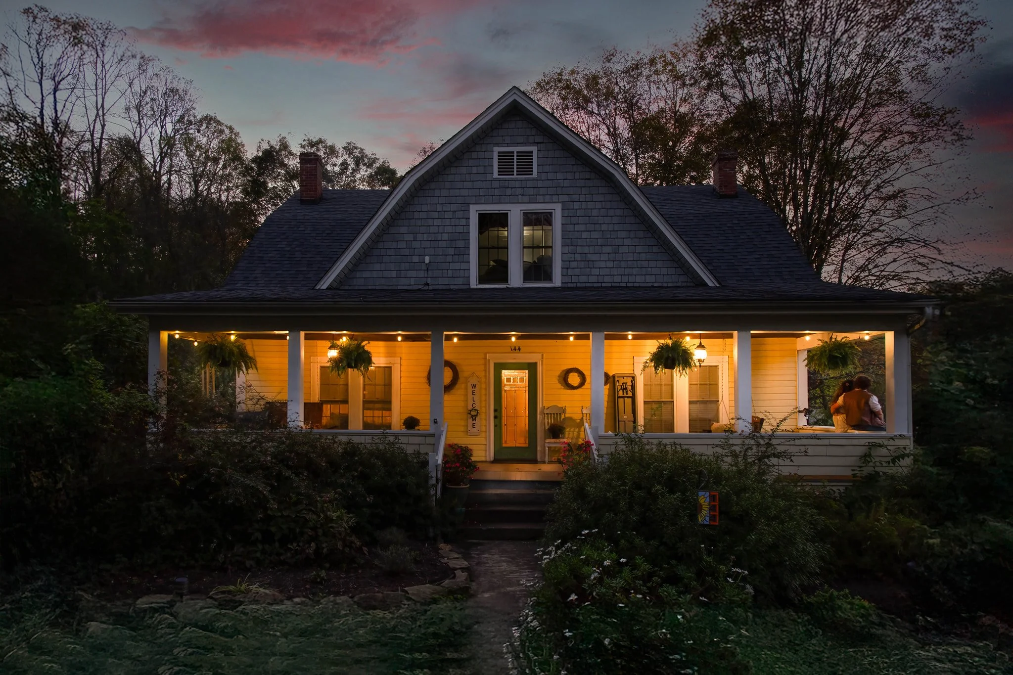The cozy Blue Note Country Inn and sweeping front porch, decorated with hanging plants and illuminated with warm string lights during dusk, surrounded by trees and a garden.