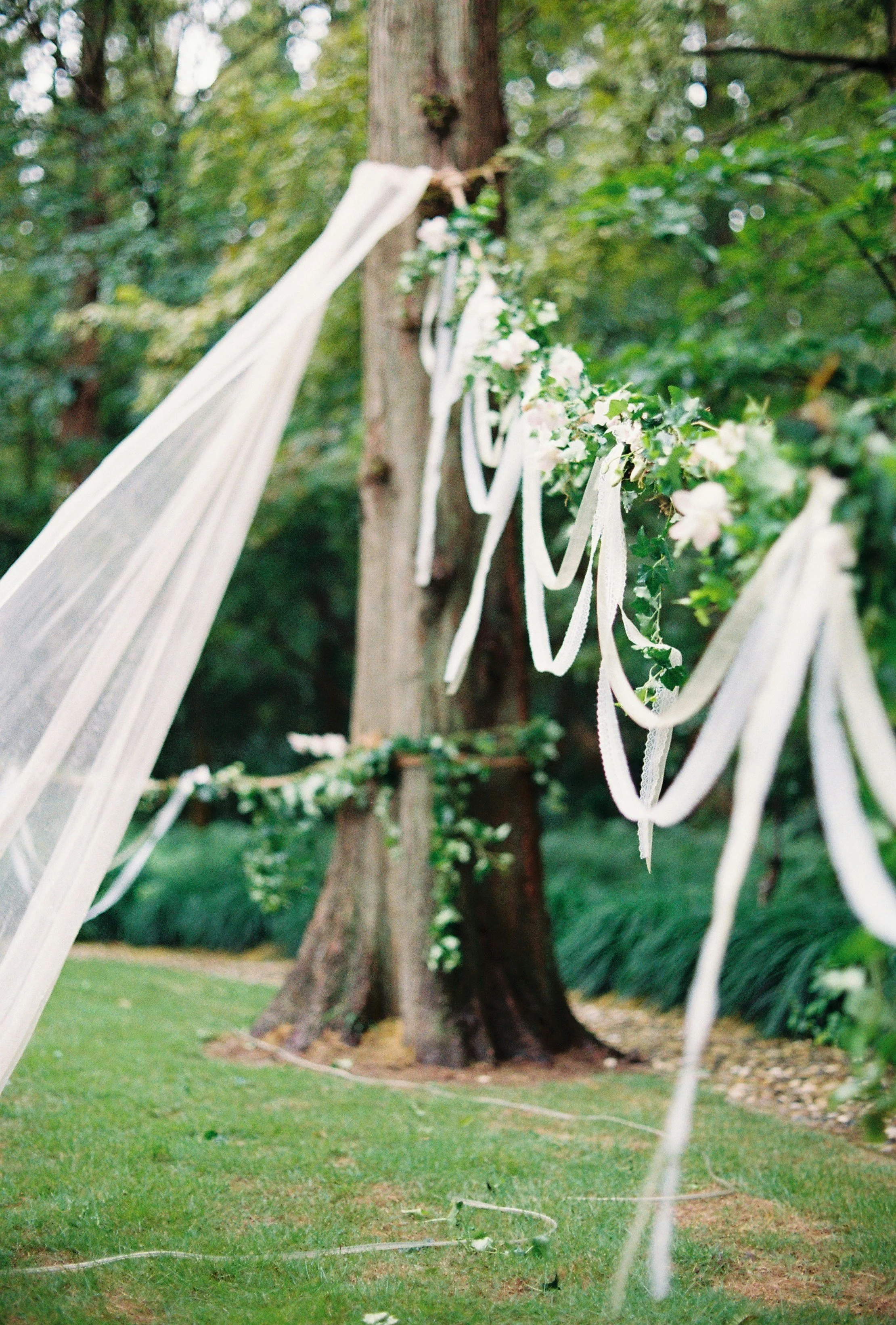 Decorative wedding arch made of tree branches, adorned with white ribbons and green foliage with white flowers, set in a wooded outdoor area.