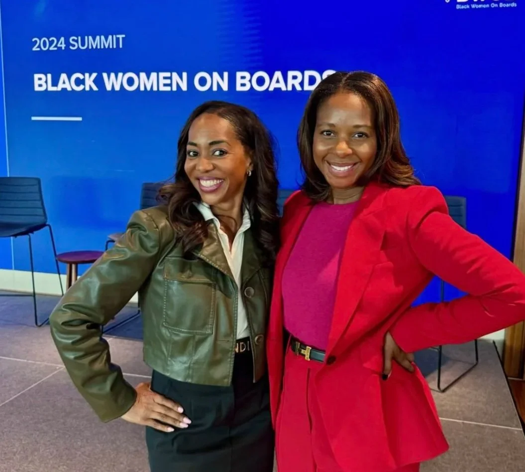 Two women smiling, standing in front of a blue backdrop that says "2024 SUMMIT BLACK WOMEN ON BOARDS". The woman on the left wears a green jacket and black pants, while the woman on the right wears a red blazer and matching skirt or pants.