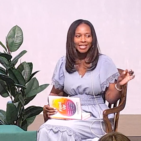A woman sitting on a wooden chair holding a book titled "Filled Up" with a colorful cover, gesturing with her right hand, wearing a light blue dress, with a large green plant nearby, and a plain white wall in the background.