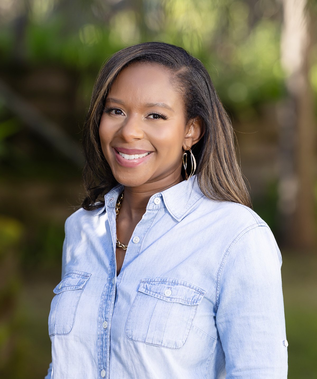 A smiling African American woman with shoulder-length dark hair and highlights, wearing a light blue button-up shirt and hoop earrings, standing outdoors with green foliage in the background.