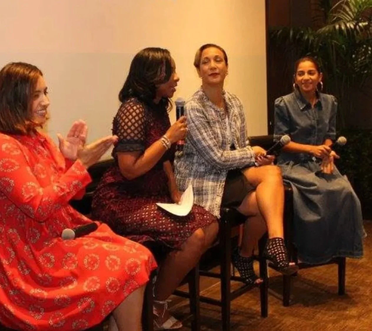 Four women sitting on chairs during a panel discussion or event. The woman on the far left is wearing a bright red dress with a floral pattern, clapping. The second woman holds a microphone, wearing a dark, patterned dress with lattice sleeves. The third woman is wearing a plaid blazer with a short skirt, holding a microphone, with her legs crossed. The woman on the far right is wearing a gray dress, holding a microphone, with a background of plants.