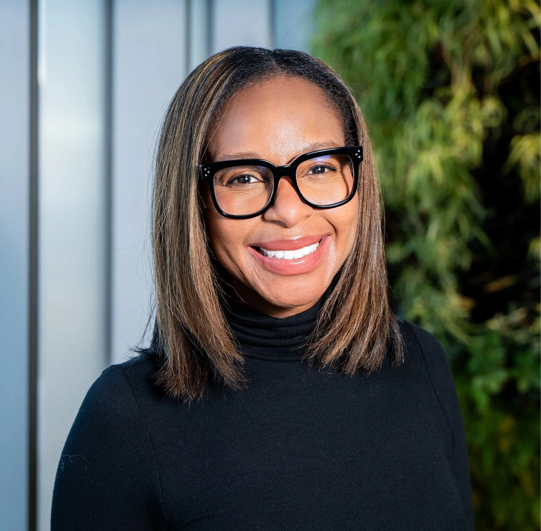 Close-up of a smiling woman with straight hair, wearing black glasses and a black turtleneck, standing outdoors with green foliage in the background.