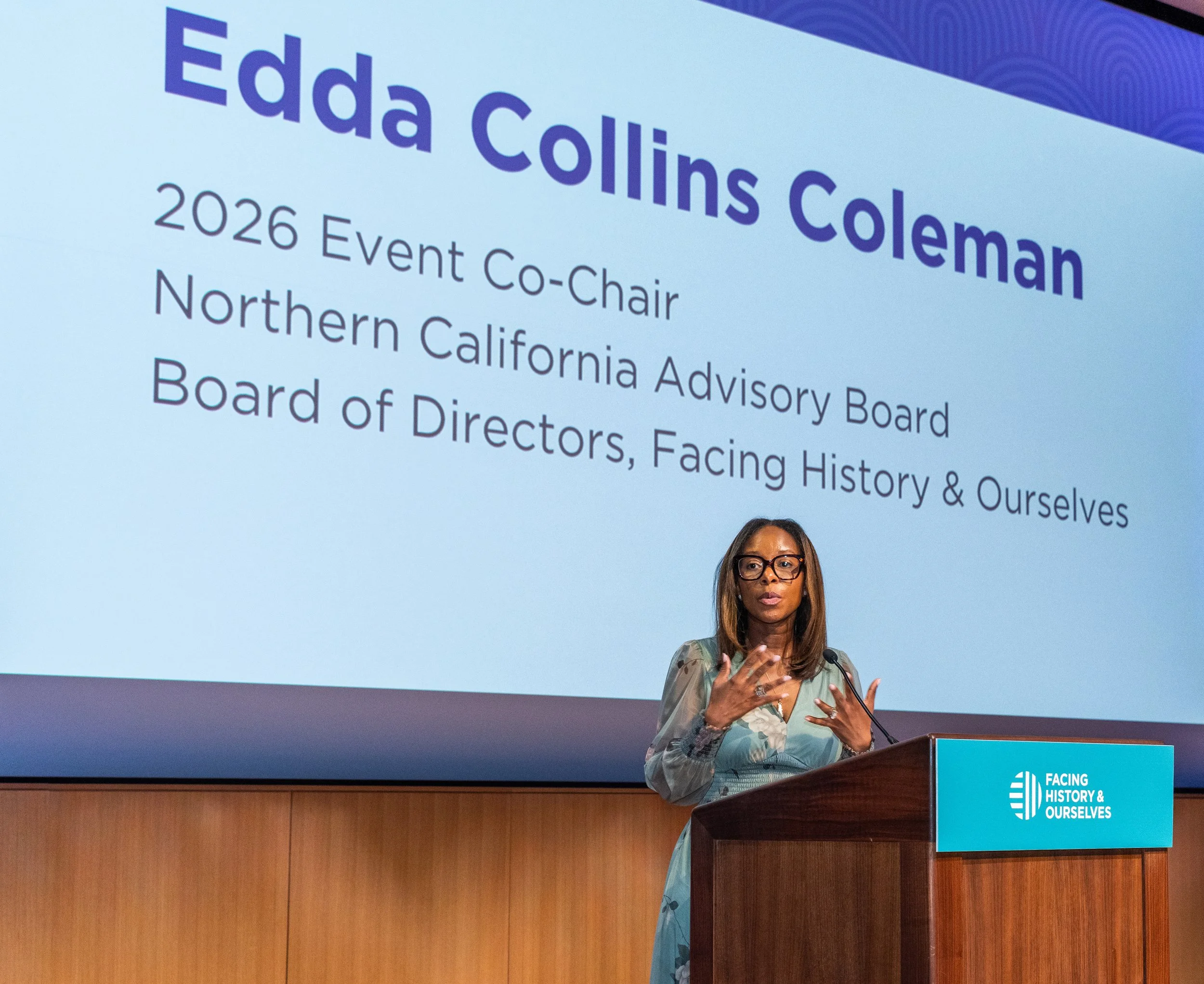 A woman with glasses speaking at a podium during an event titled 'Facing History & Ourselves,' with a large screen behind her displaying the same event title and additional information about the event.