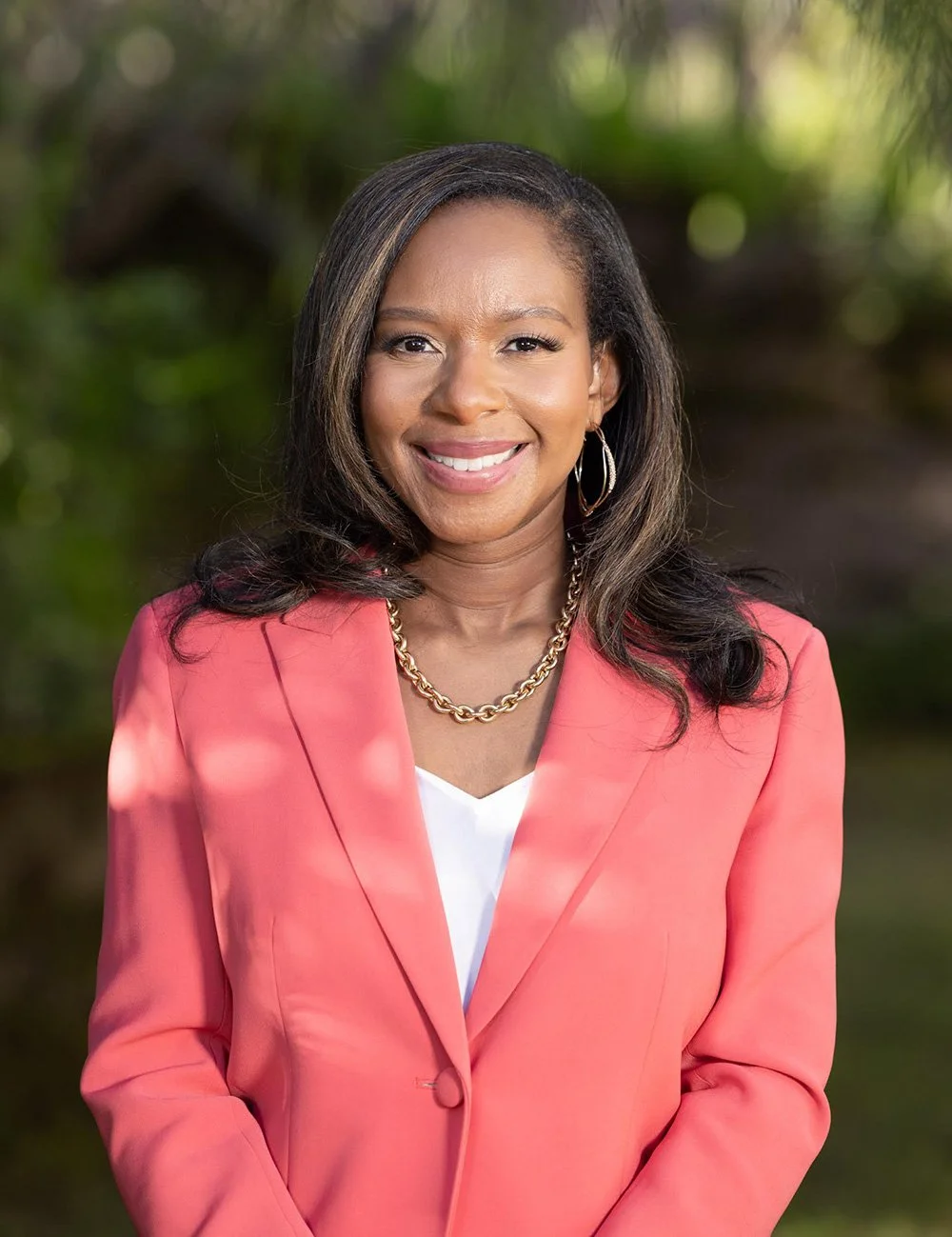 A woman with medium-length black hair, smiling, wearing a coral blazer, white top, gold chain necklace, and hoop earrings, standing outdoors with blurred green foliage in the background.