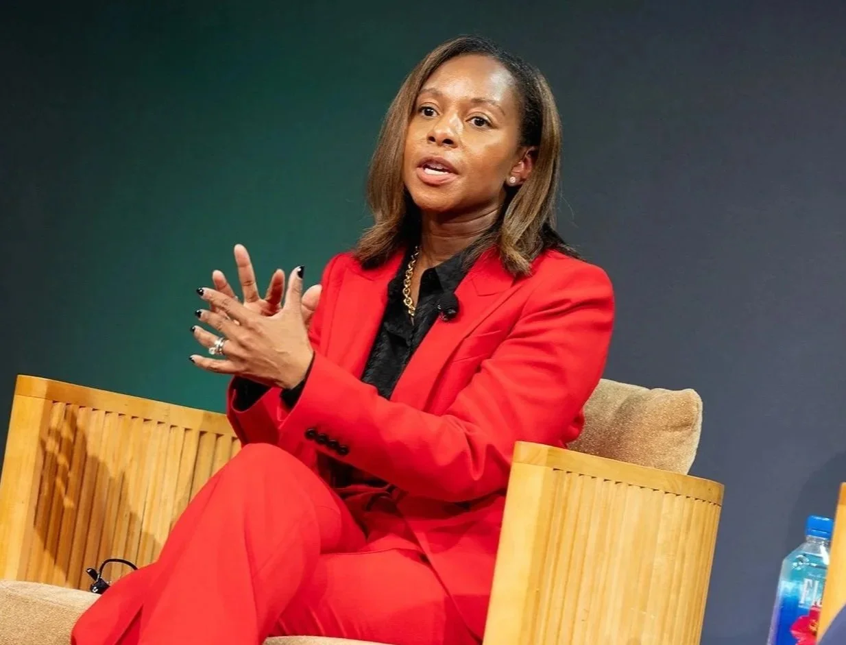 Woman sitting in a wooden chair, wearing a red suit and speaking, with a dark background and water bottle nearby.