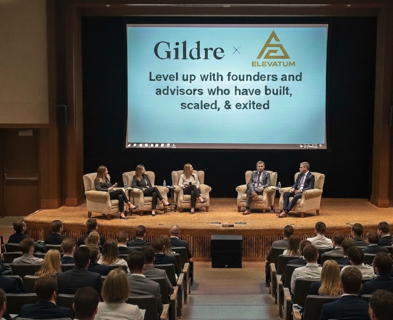 A panel discussion with five speakers seated on stage in front of an audience, with a large screen behind them displaying the logos of Gildre and Elevatum and the text, 'Level up with founders and advisors who have built, scaled, & exited.'