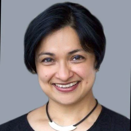 Portrait of a smiling woman with short black hair, wearing a black top and a necklace with a crescent pendant, against a gray background.