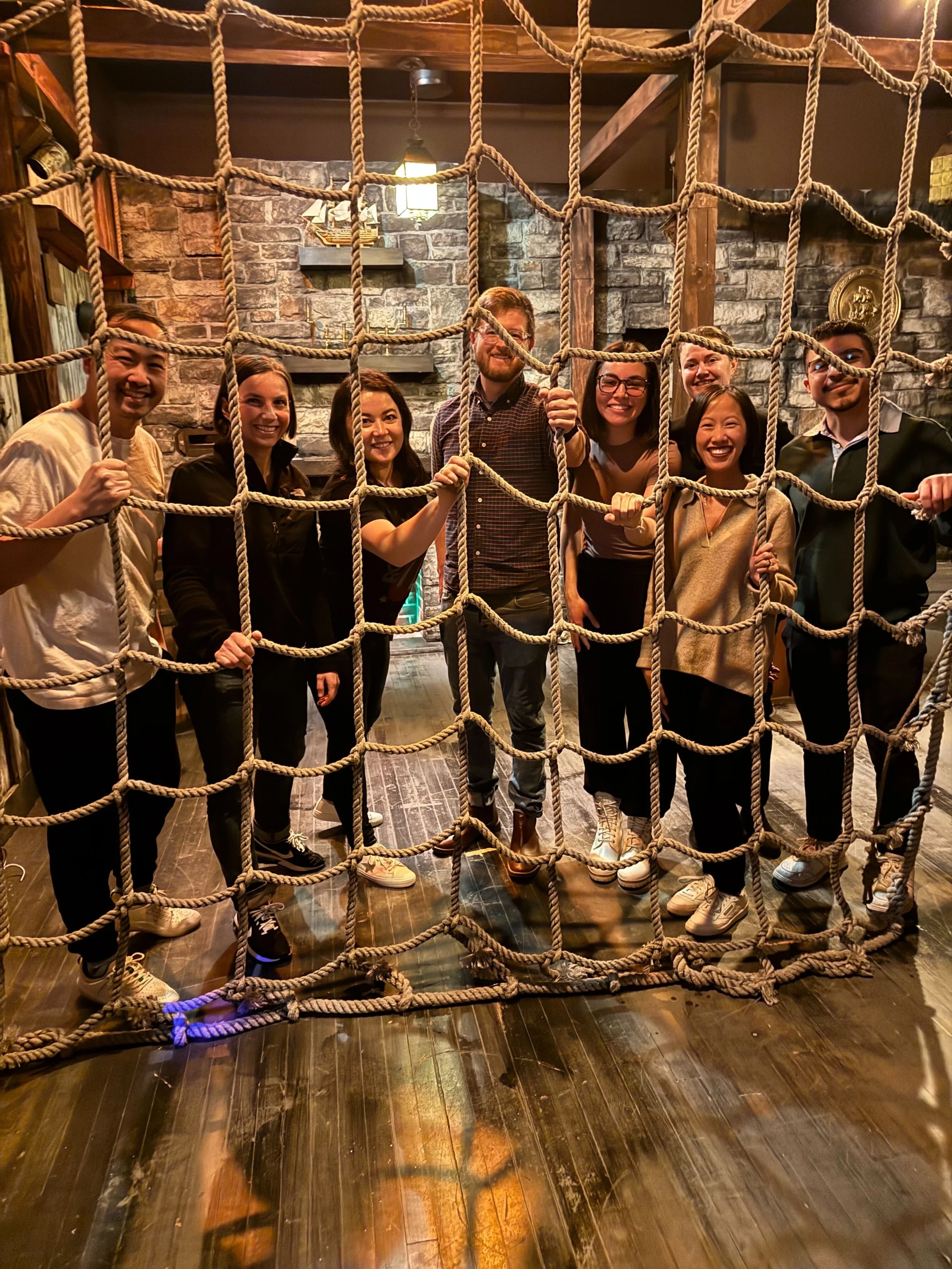 A group of eight people standing behind a large rope net indoors. The setting appears to be rustic with stone walls and wooden beams, suggesting a themed or escape room environment. They are smiling and appear to be enjoying the experience.