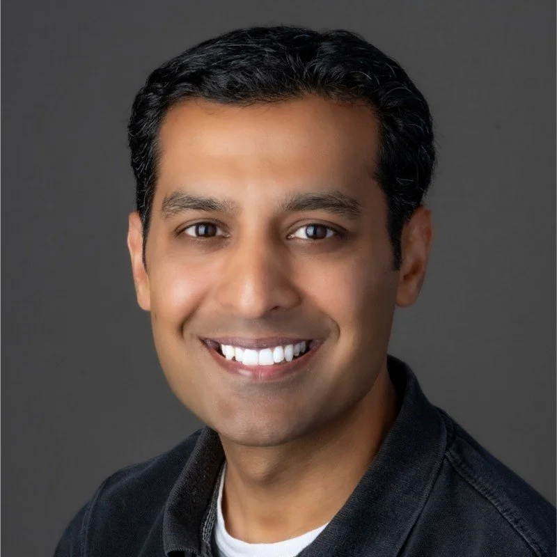Smiling person with short dark hair wearing a black shirt on a gray background.