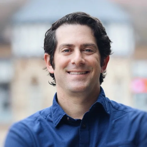 Man with dark hair smiling, wearing a blue shirt, blurred background.