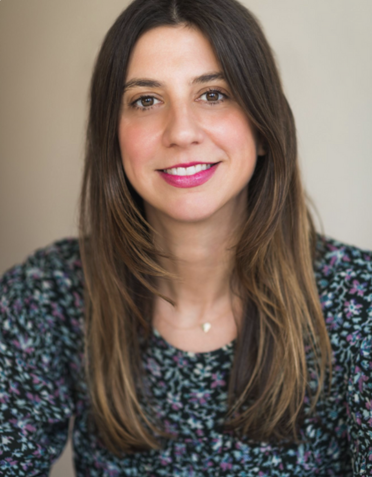 A woman with long brown hair, smiling with pink lipstick, wearing a black and purple floral top and a delicate necklace, against a neutral background.