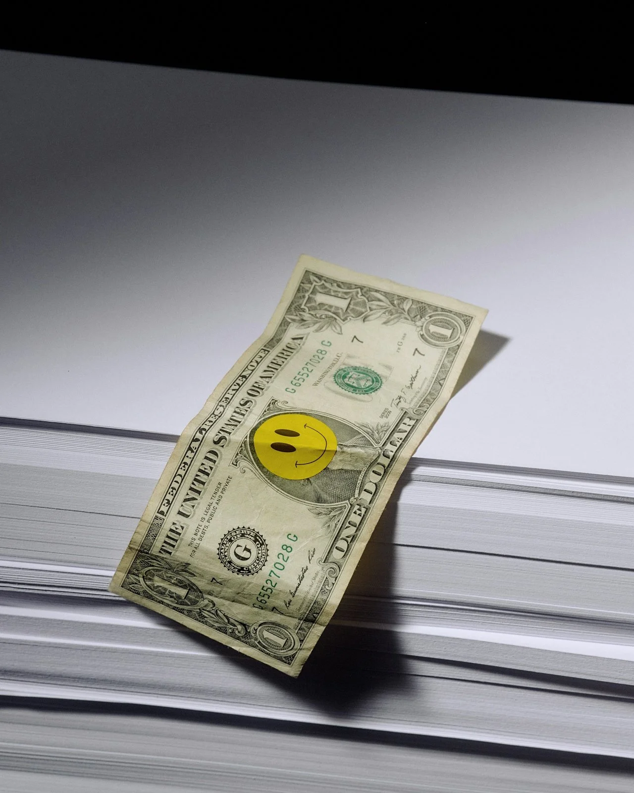 One dollar bill with a yellow smiley face sticker on it, placed on top of a large stack of papers or documents.
