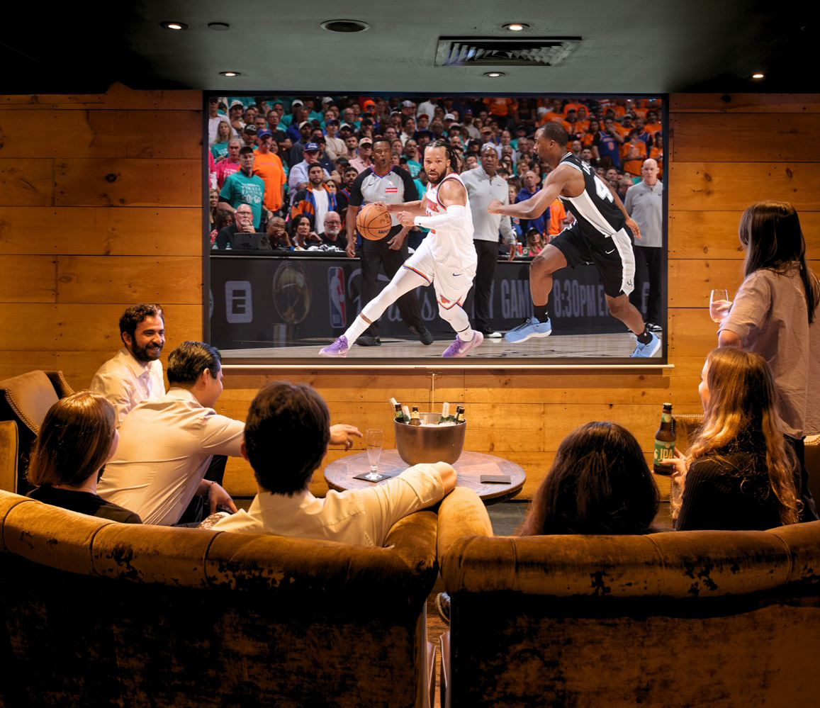 A group of friends gathered on a curved velvet sofa in BLACKBARN's wood-paneled private room, watching a basketball game on a large screen with a chilled bottle service pail on the table