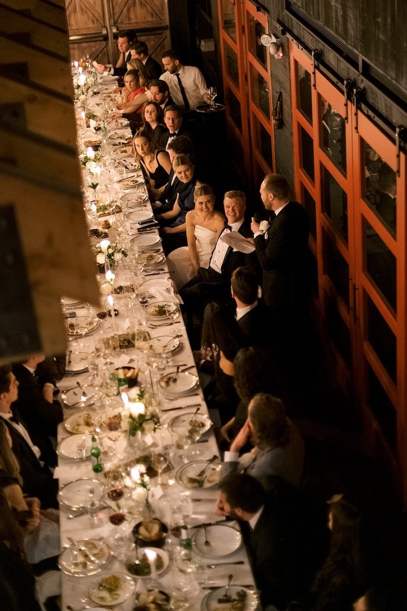 Aerial view of a long banquet table at a formal event with guests seated. The table is elegantly set with plates and glassware, and candles are lit along the center. A person is standing and speaking to the seated guests, including a couple in formal