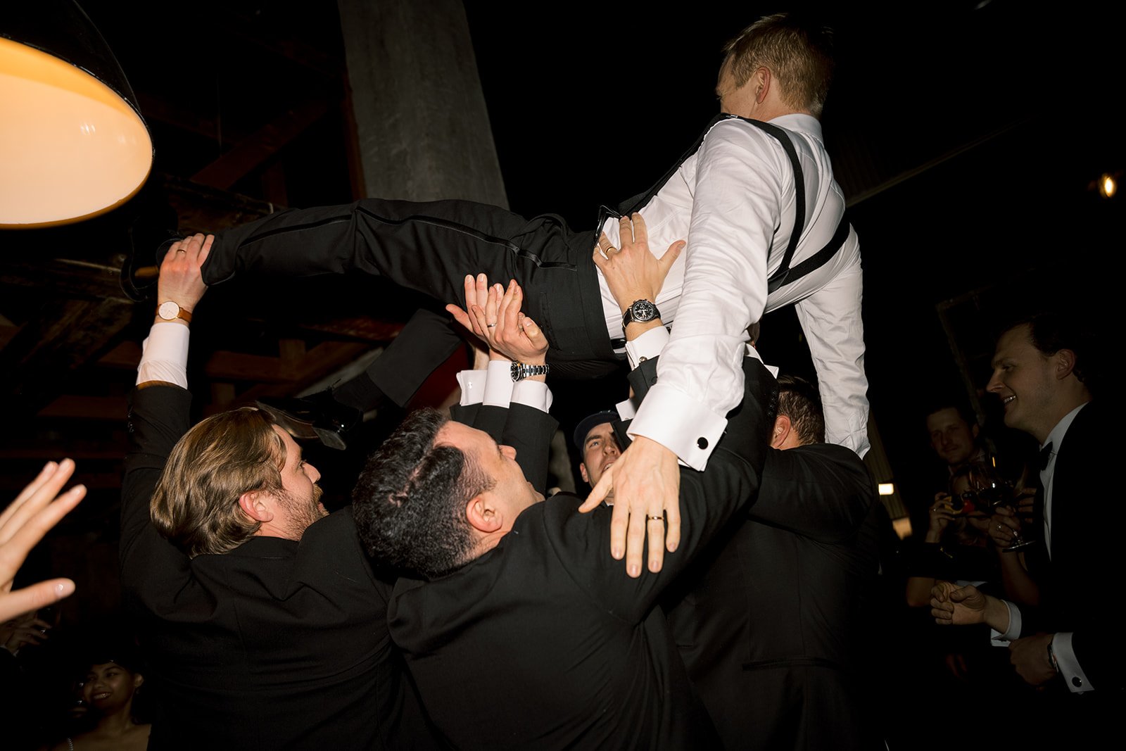 The horah–Group of men in suits celebrating, lifting another man in a white shirt and suspenders in the air during a party.