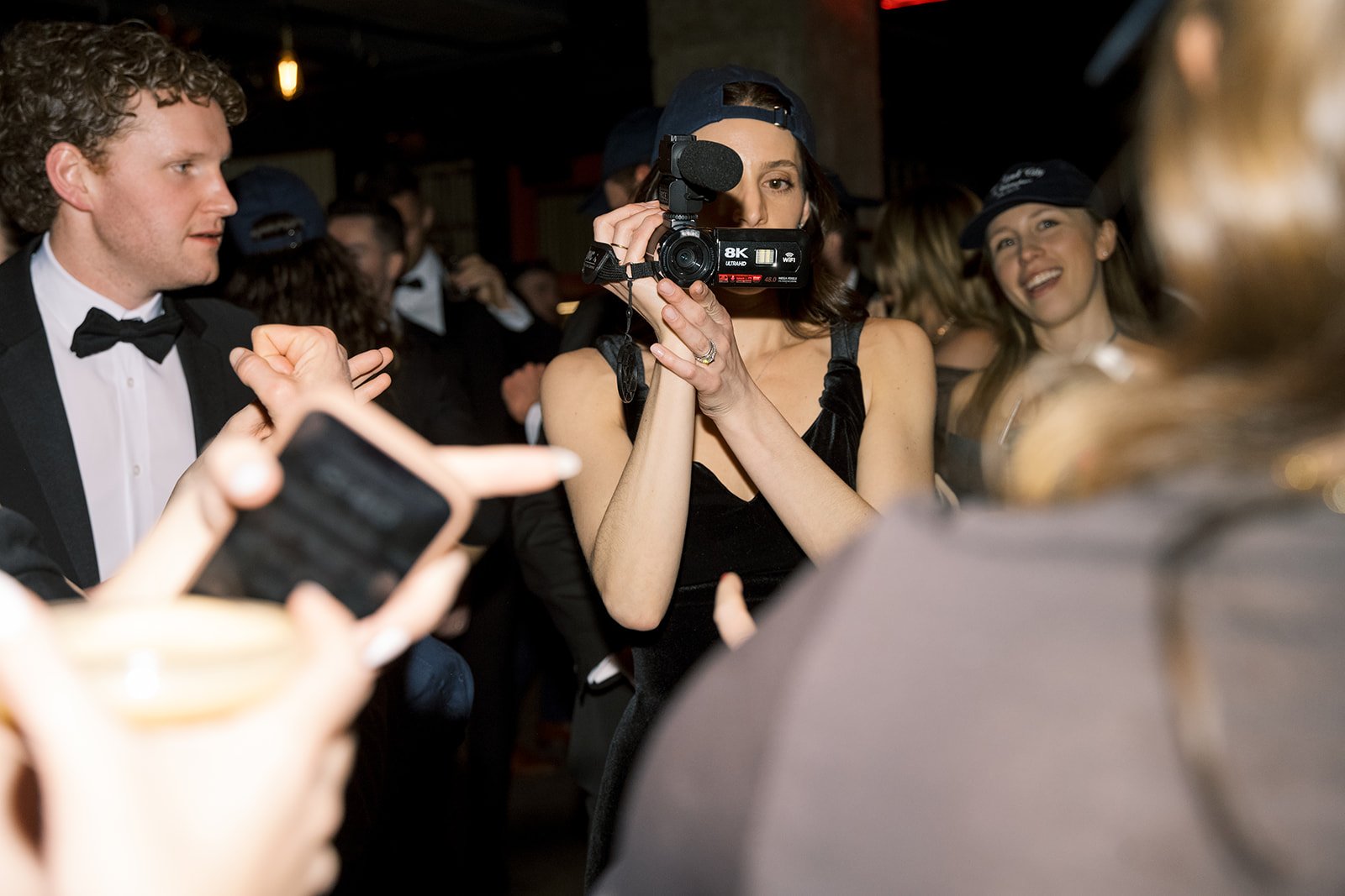 A woman in a black dress holding a video camera at a party with elegantly dressed people in the background.