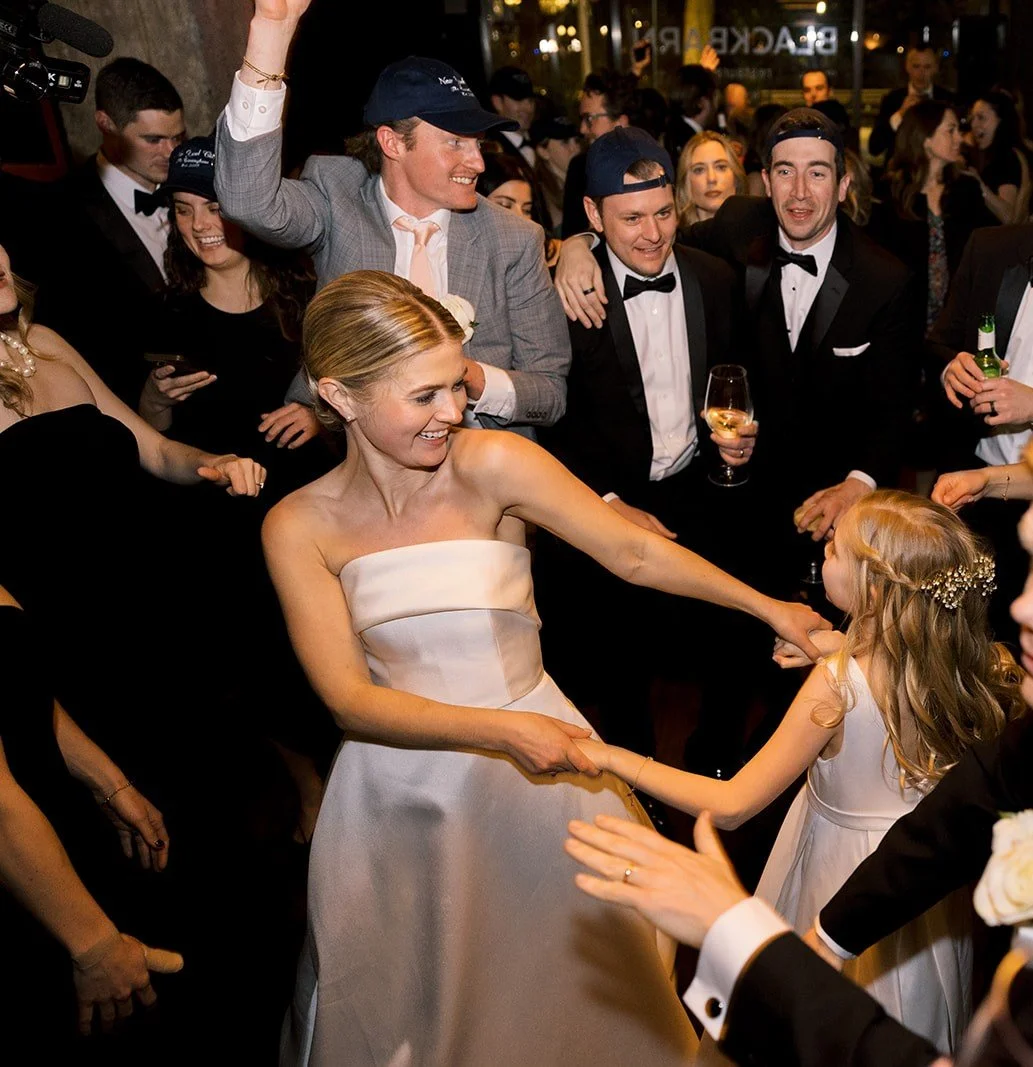 A wedding reception with a bride dancing with a young girl, surrounded by guests in formal wear, some wearing hats, enjoying the celebration.