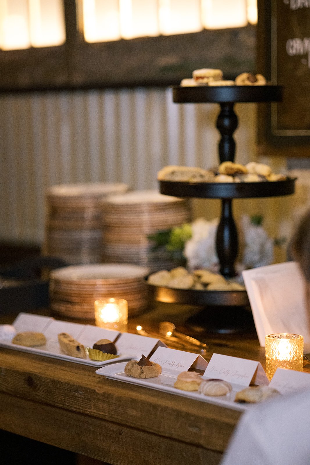 Dessert table with tiered stand holding cookies, plates stacked in the background, candles, and name cards for each dessert on a wooden table.