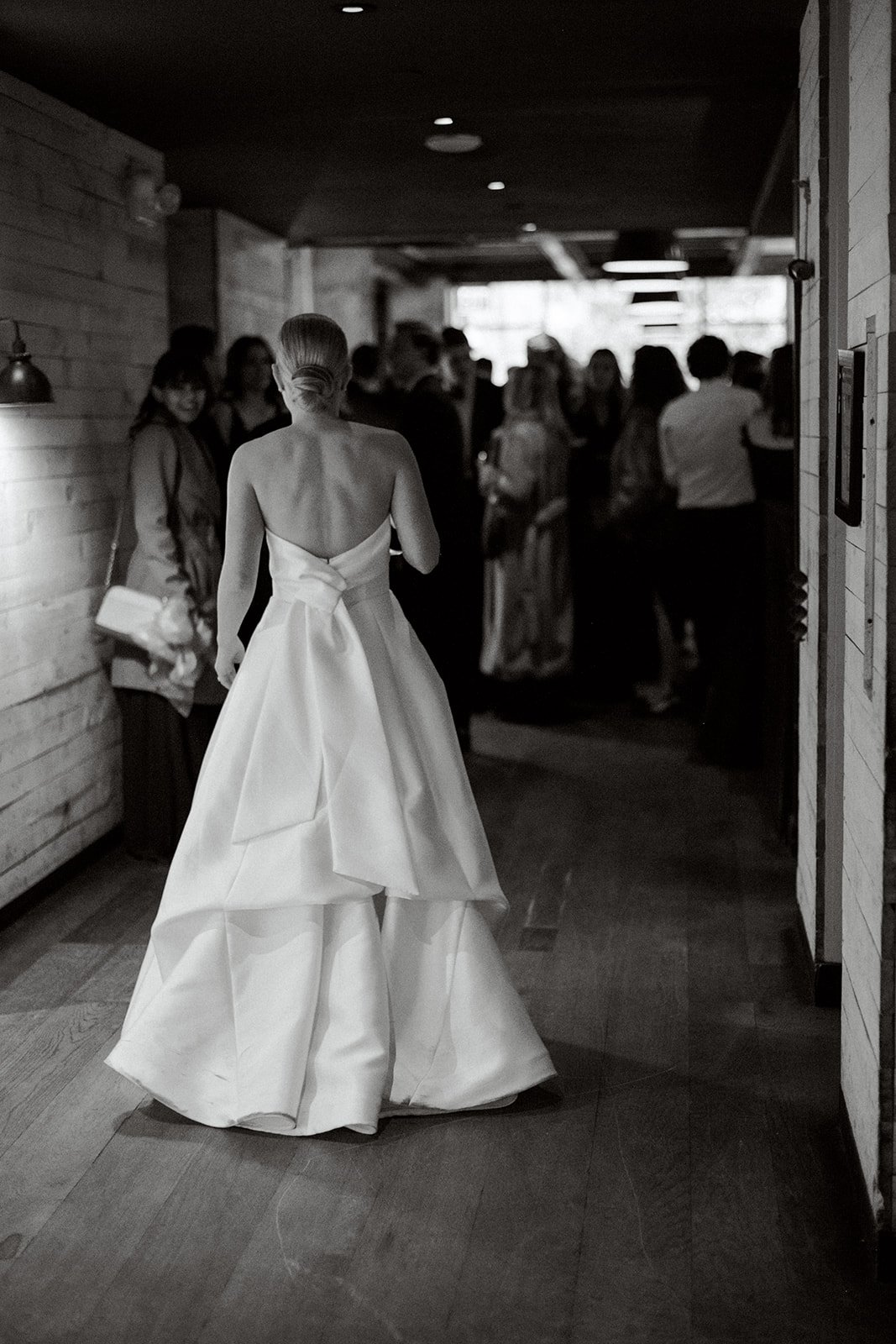 Black and white photo of a person in a wedding dress walking down a hallway towards a group of people.