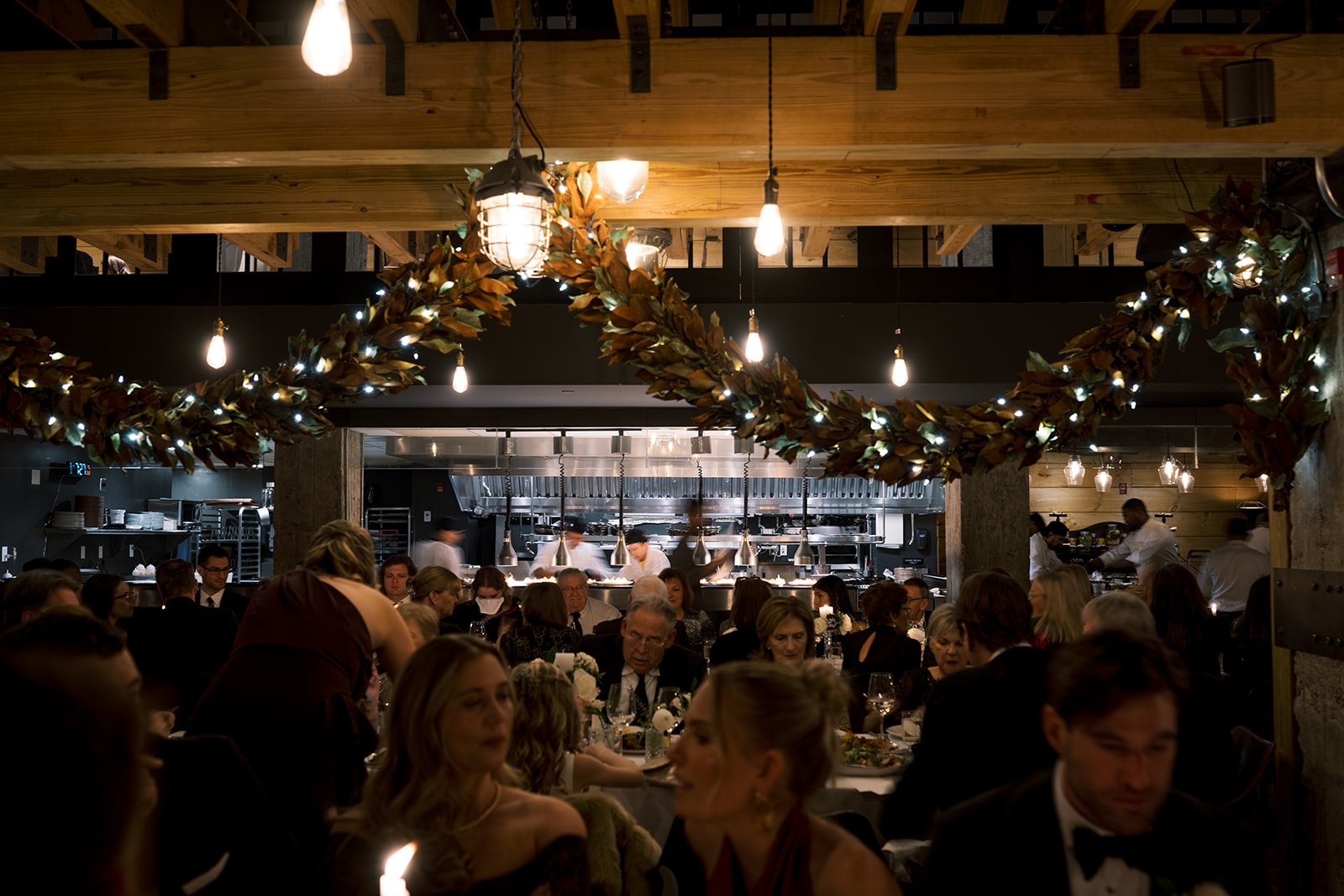 Elegant dining room with guests seated at tables, open kitchen in the background, warm lighting and decorative garlands.