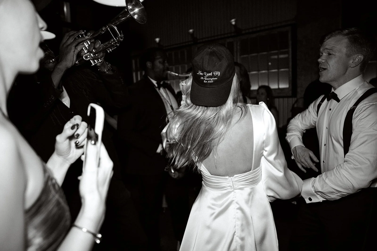 A black and white image of people dancing at a party. A woman in a satin dress and baseball cap is in the foreground, while a man in formal attire dances nearby. A trumpet player is performing in the background and another woman is holding a smartpho