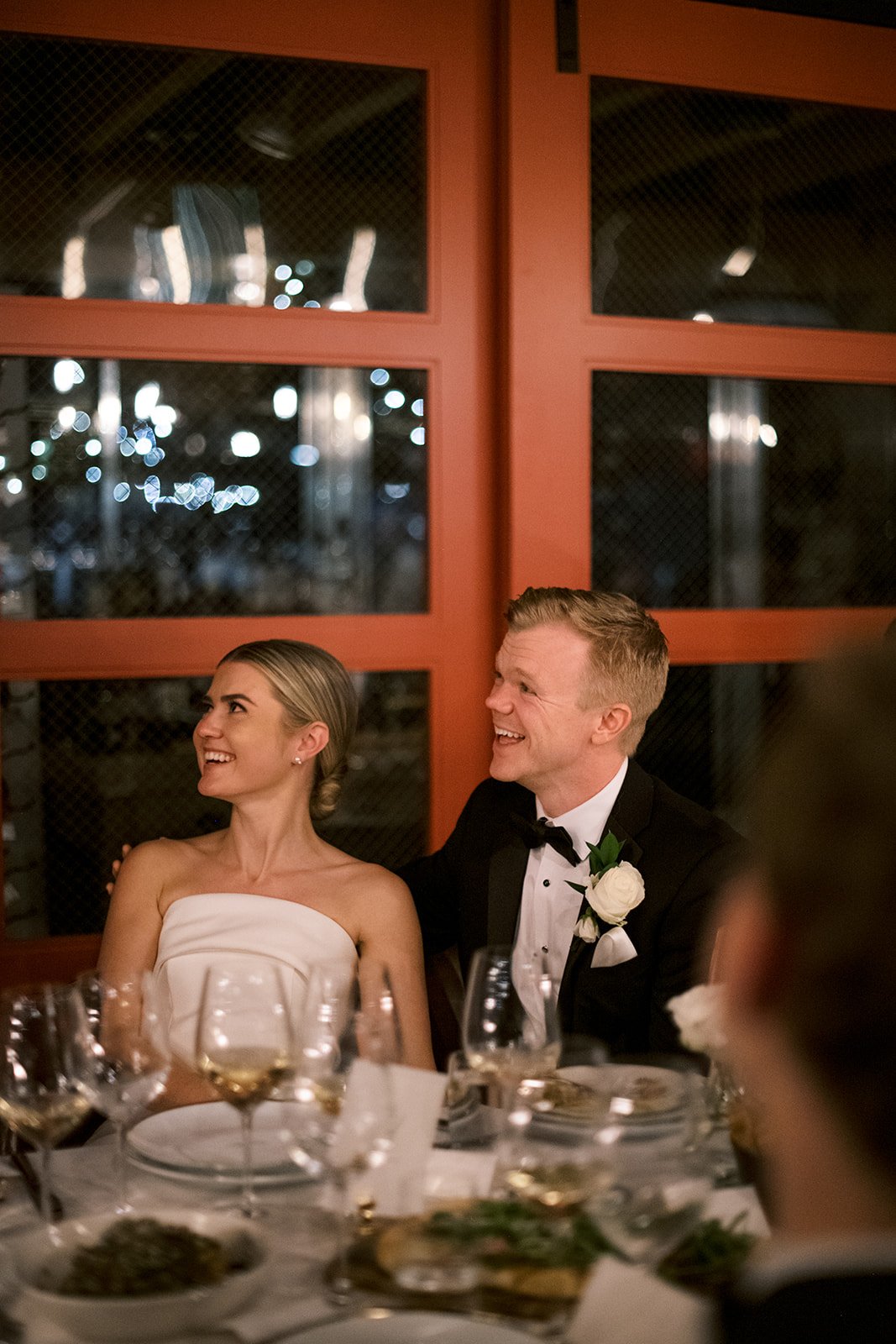 A bride and groom smiling at their wedding reception, seated at a table with wine glasses and plates, with a red backdrop.