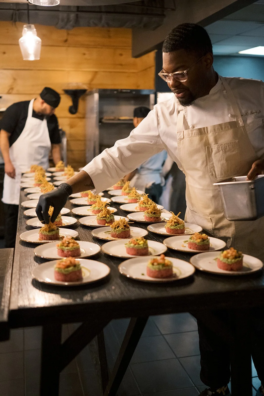 Chefs preparing multiple plates in a professional kitchen, with dishes featuring layered food garnished artistically. One chef is arranging the ingredients wearing a glove, while another chef works in the background. The scene is set in a warmly lit