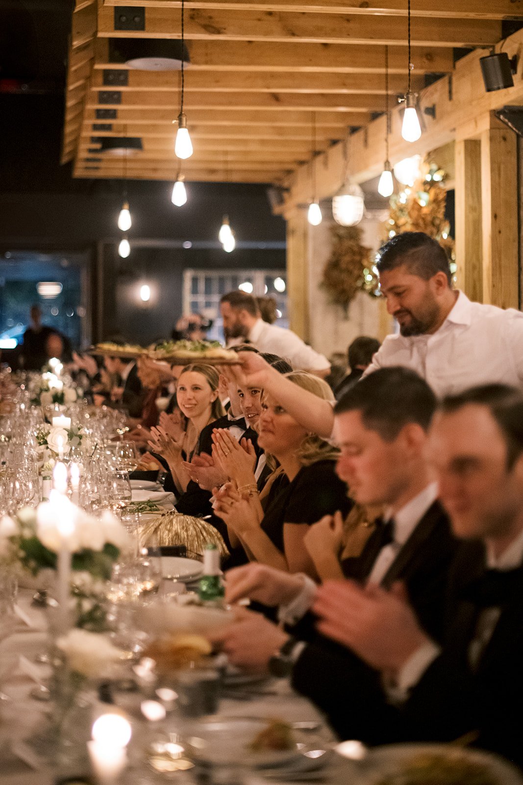 People seated at a long table in a dimly lit restaurant, clapping and enjoying a formal dinner, with a waiter serving dishes.