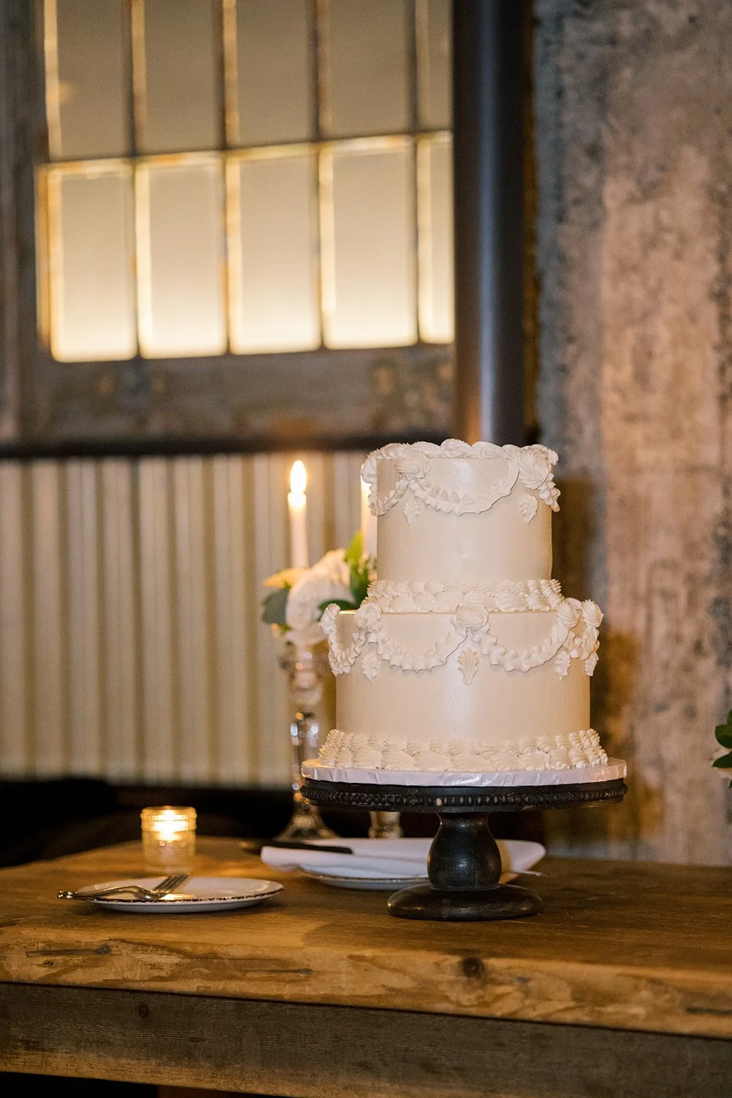 A decorative two-tiered white wedding cake on a wooden table with a candle and plates nearby.
