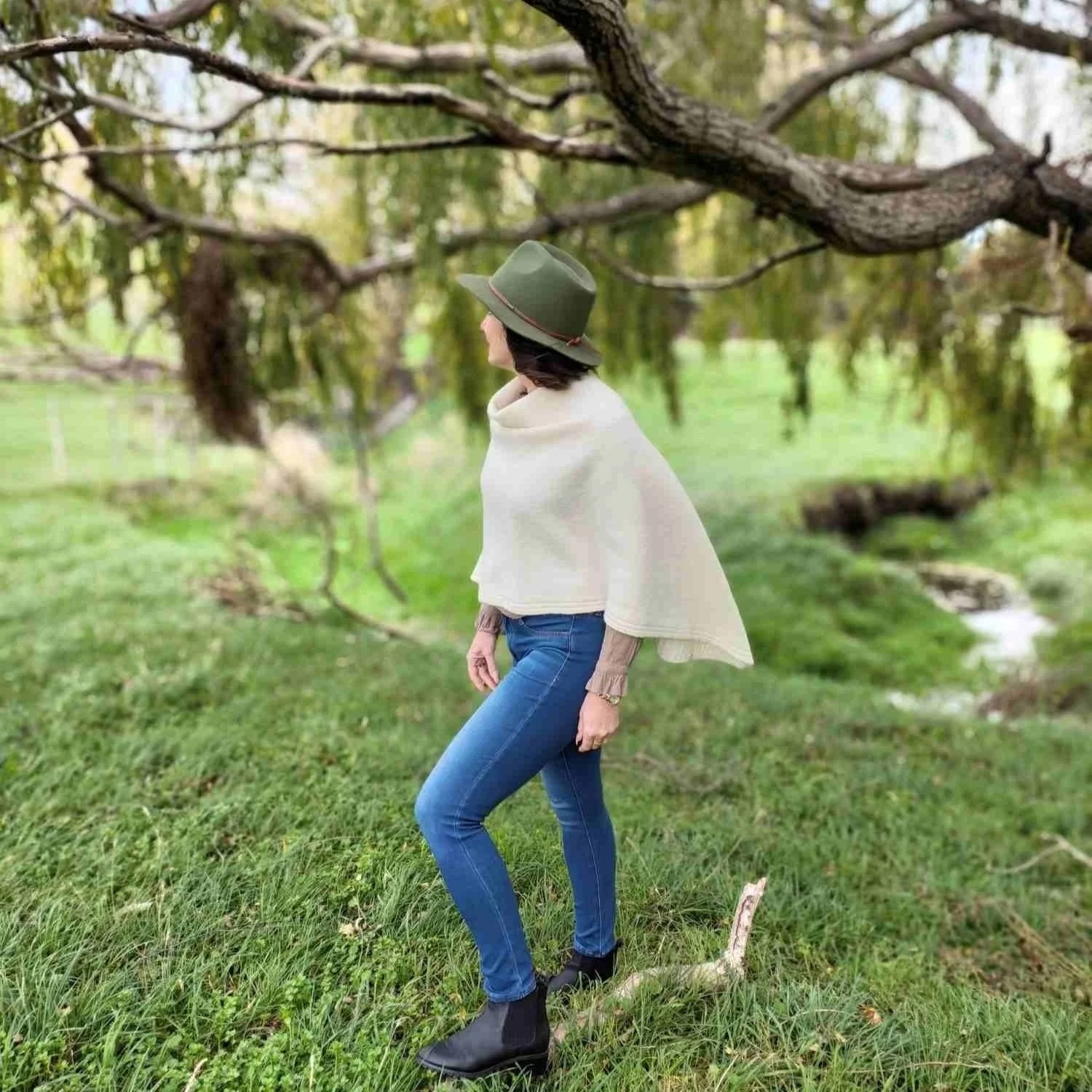 A woman standing outdoors on grass near a tree with hanging branches, wearing a green hat, Bohepe cream poncho, brown long-sleeve shirt, blue jeans, and black ankle boots at Ataahua Farm on Banks Peninsula