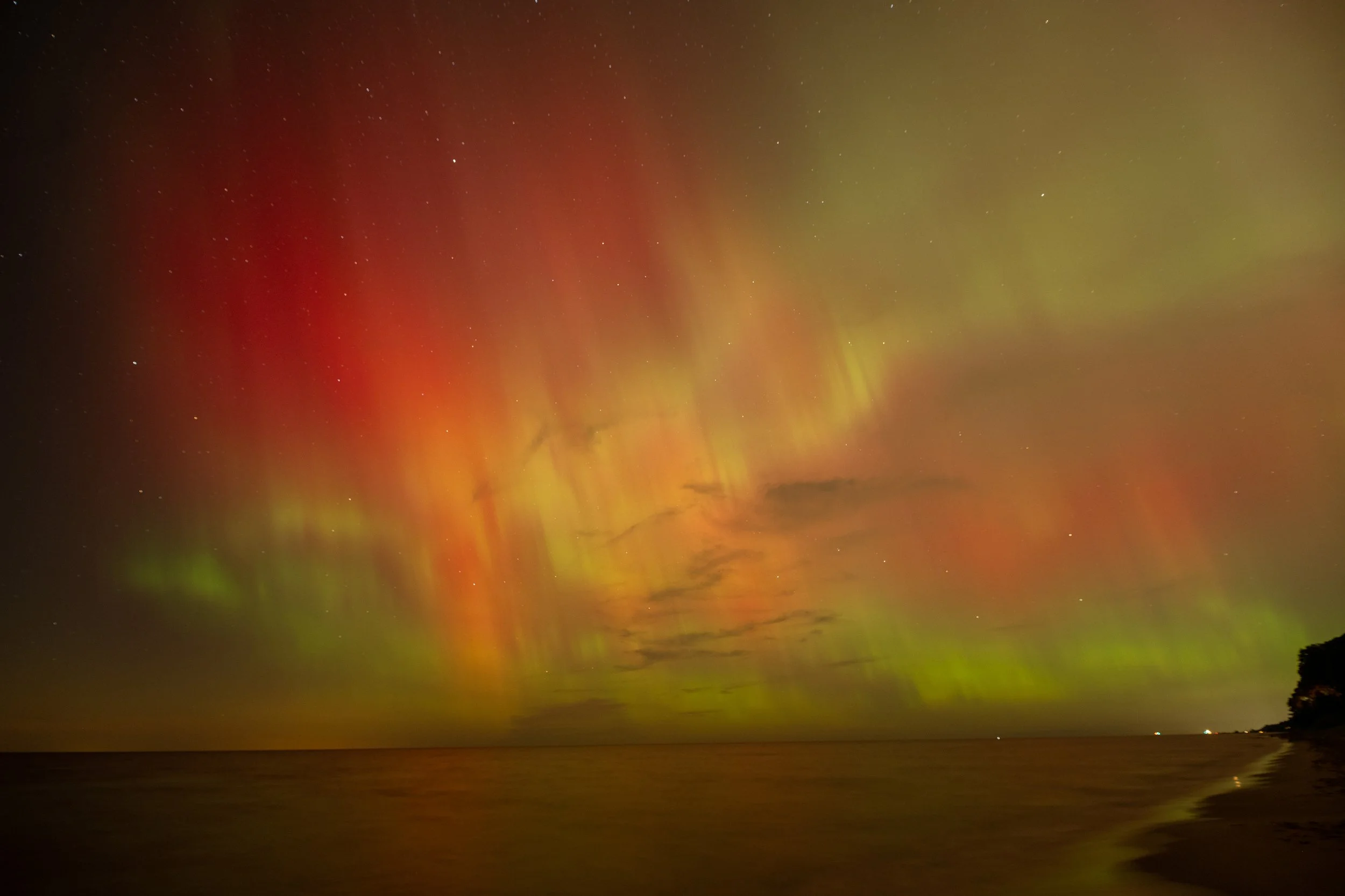 Vibrant aurora borealis with red, orange, and green colors over a calm sea, visible under a starry night sky.