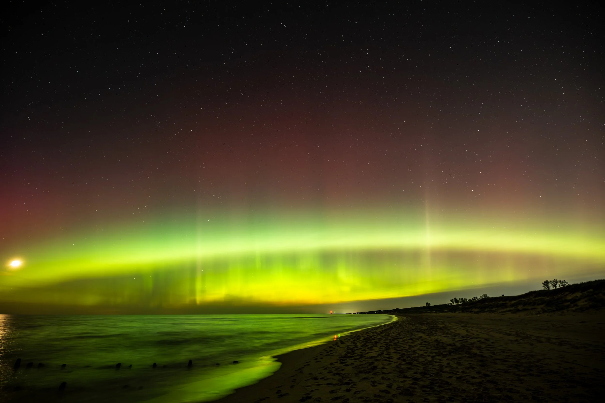 Northern Lights over a scenic beach at night, with green and red auroras visible in the sky and reflection on the water.