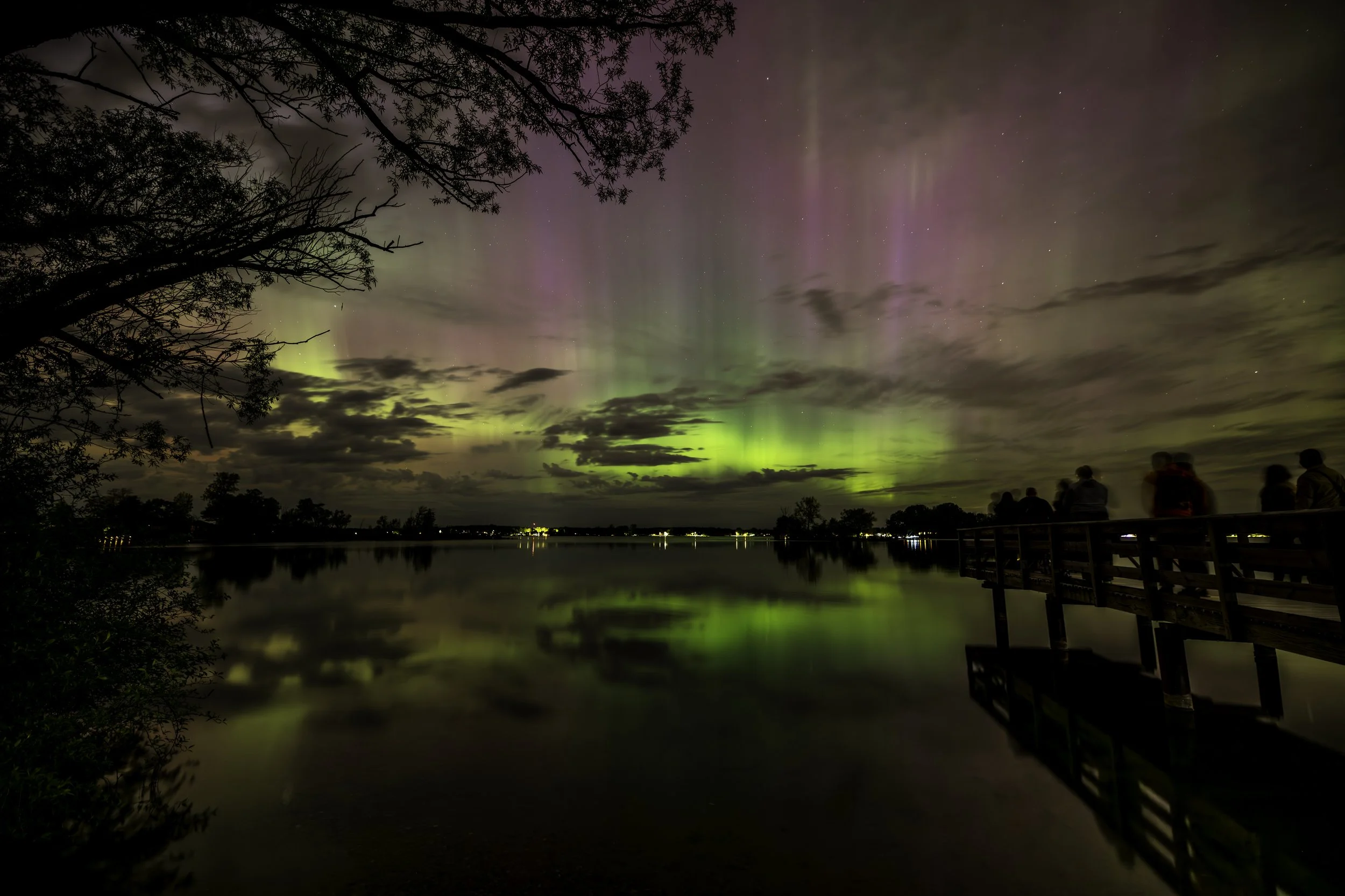 Northern lights over a lake with silhouettes of people on a dock at night.