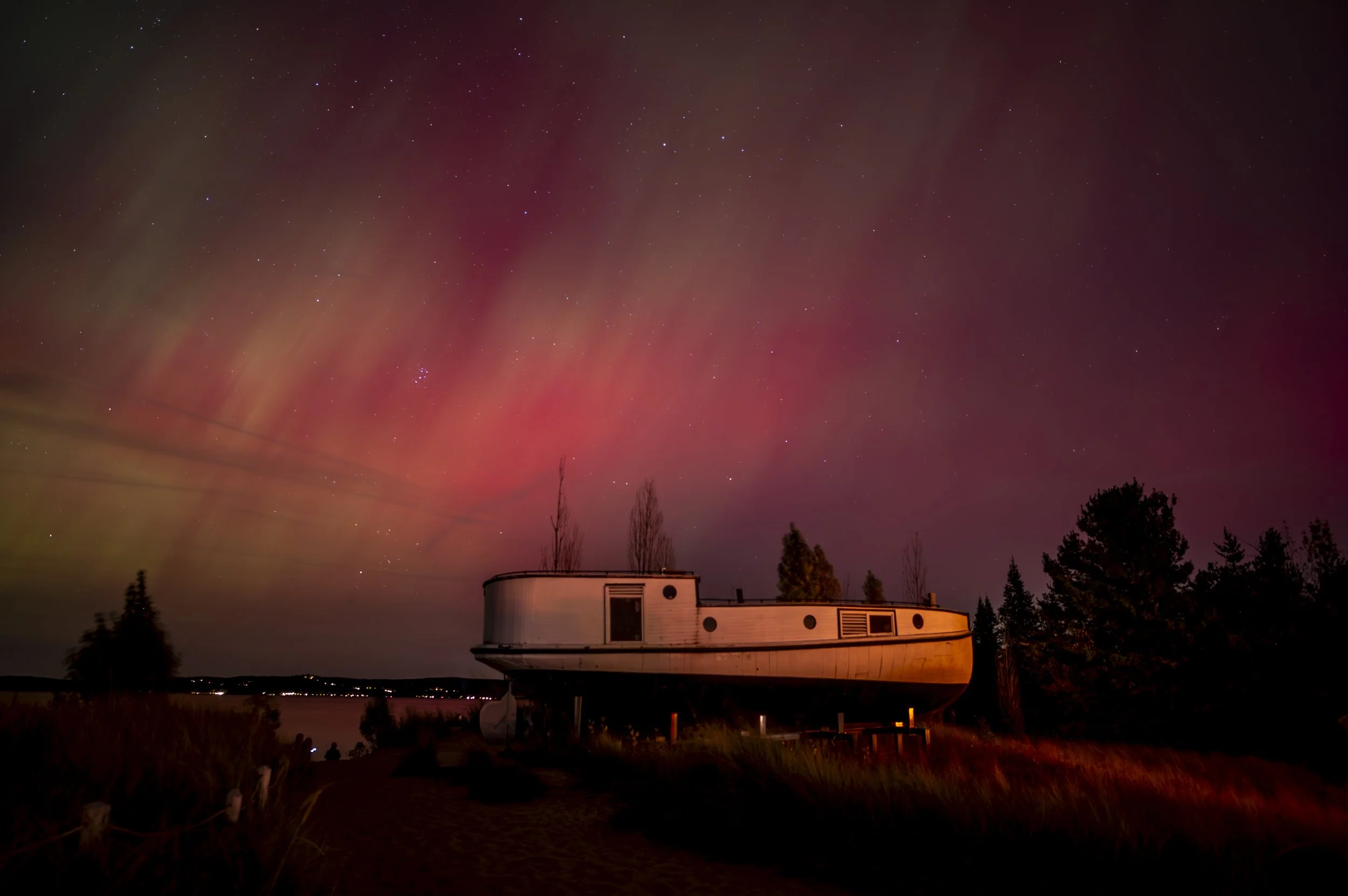 A boat-like structure on land under a colorful aurora-filled night sky with red and green hues and scattered stars.