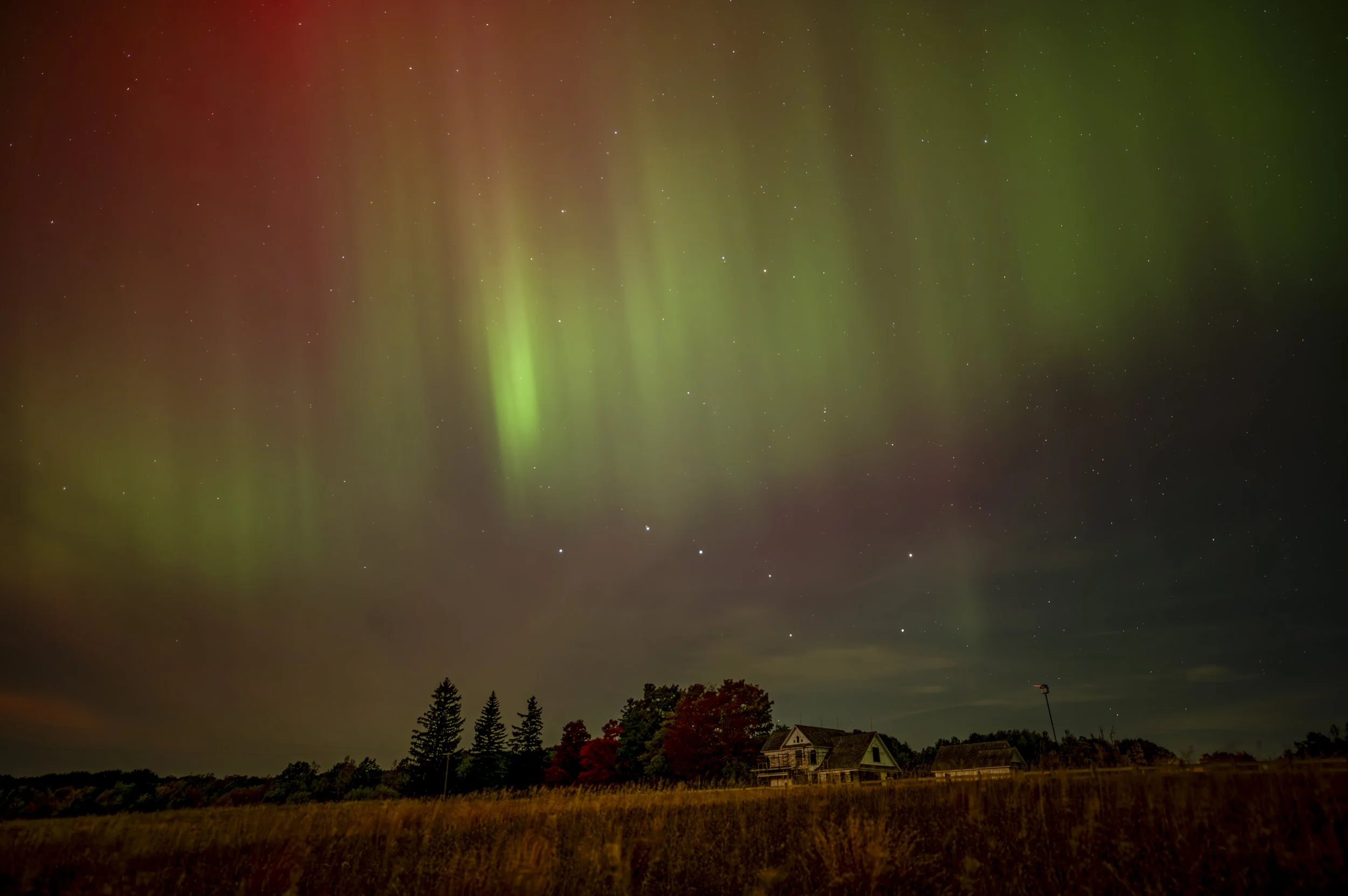 Colorful aurora borealis over a rural landscape with a house and trees.