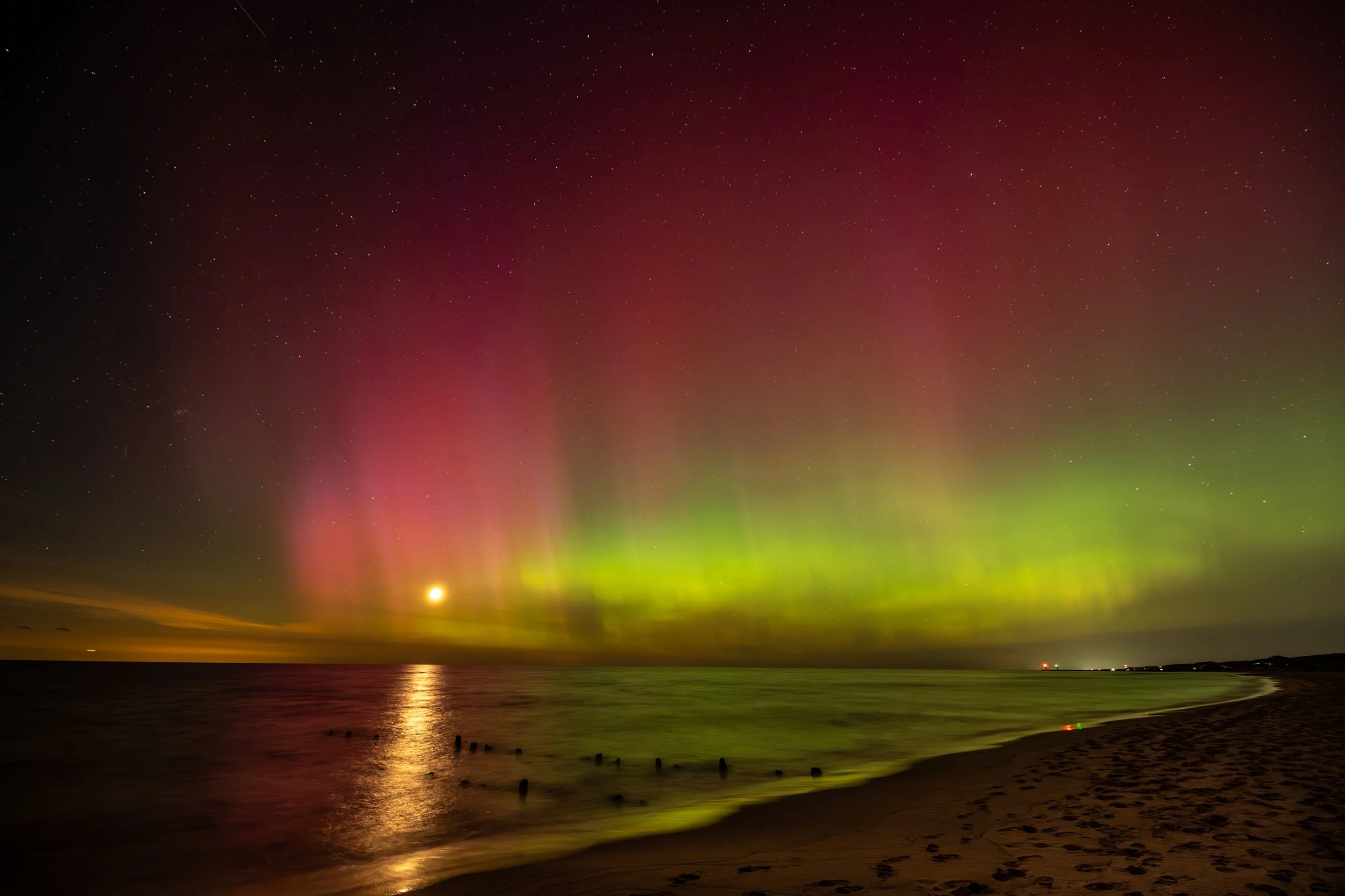 A brilliant green aurora arc spans the horizon over Lake Michigan at Oval Beach in Saugatuck, Michigan, casting green light on the waves, old pier pilings and footprint-covered sand, May 2024.