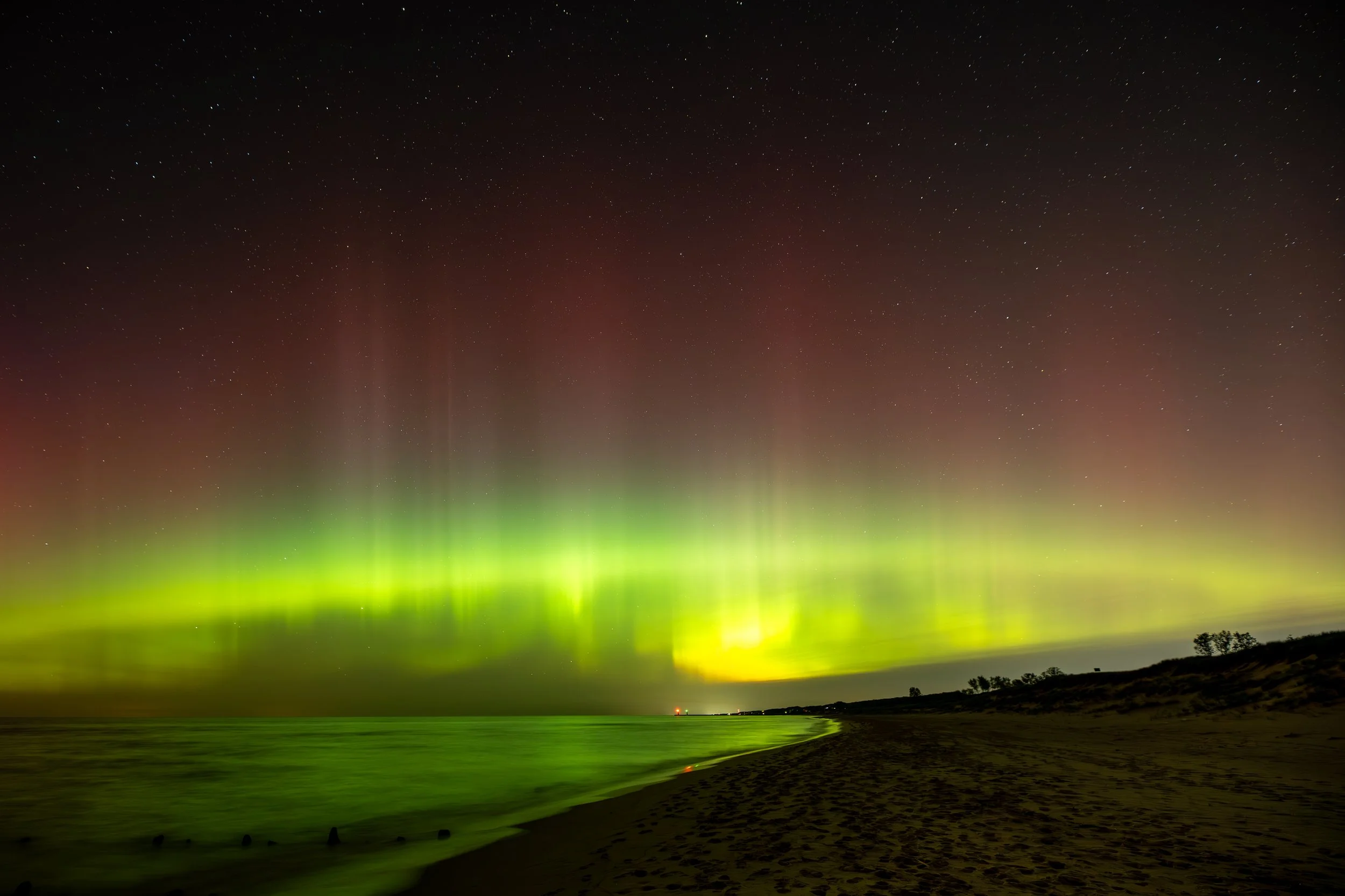 Vibrant aurora borealis over a beach at night, with green and red lights reflecting on the water.