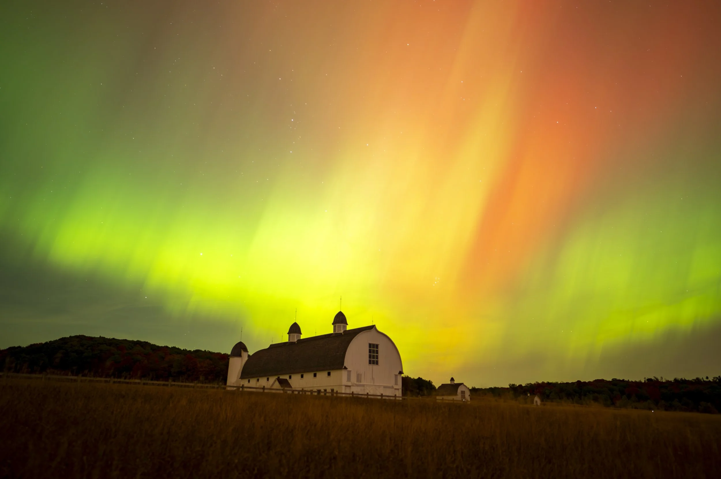 Northern lights over a white barn at night with a grassy field in the foreground and trees in the background.