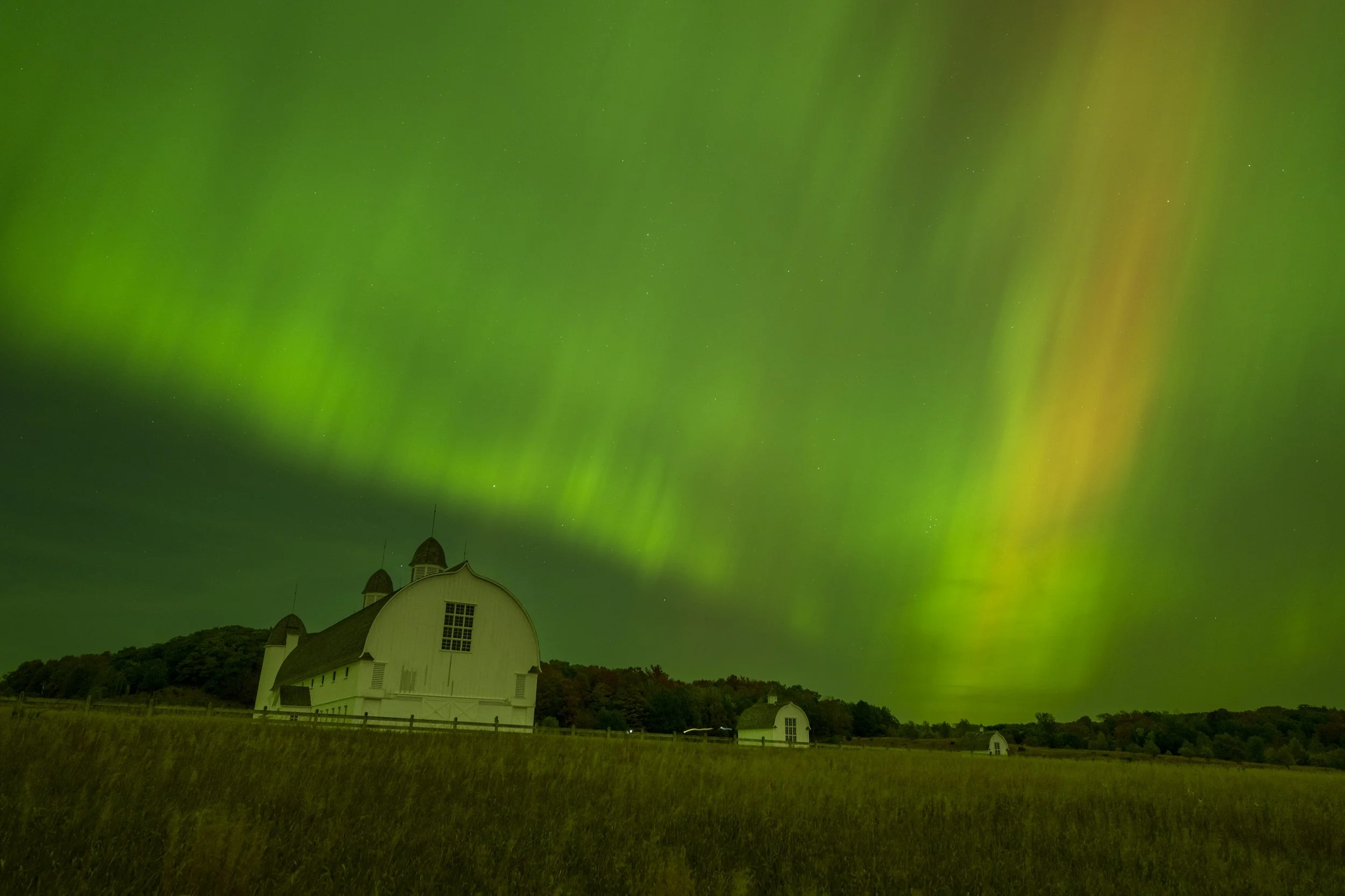 Northern lights over a white barn in a rural field at night.