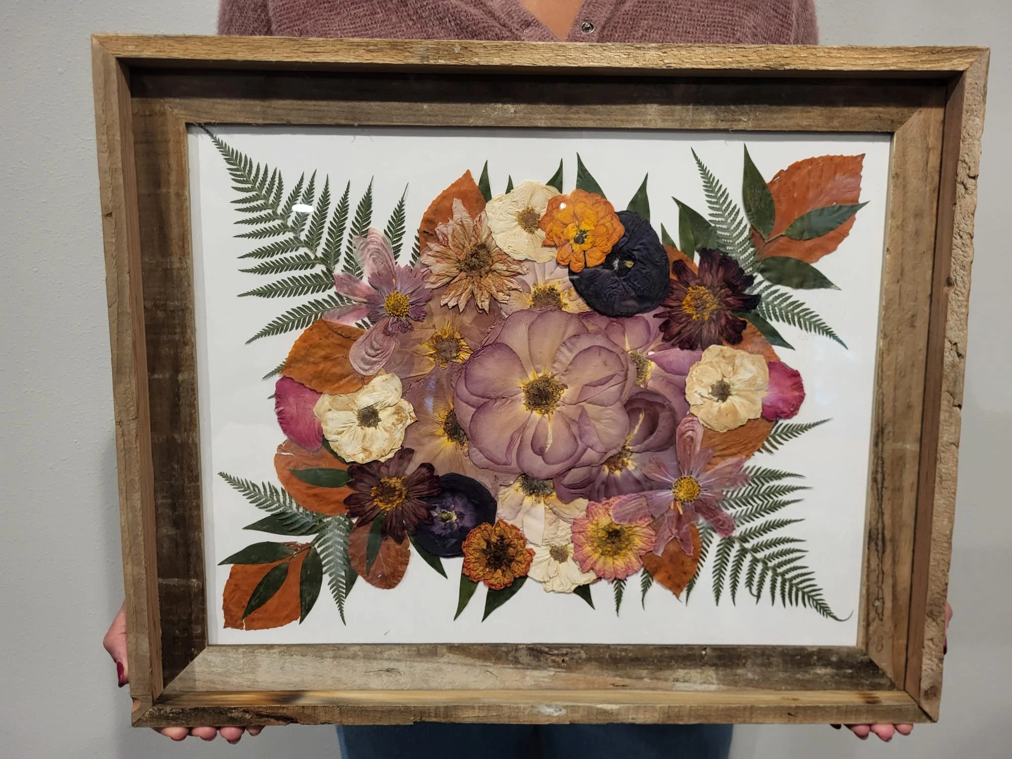 A framed pressed flower arrangement featuring various colorful dried flowers and green fern leaves.