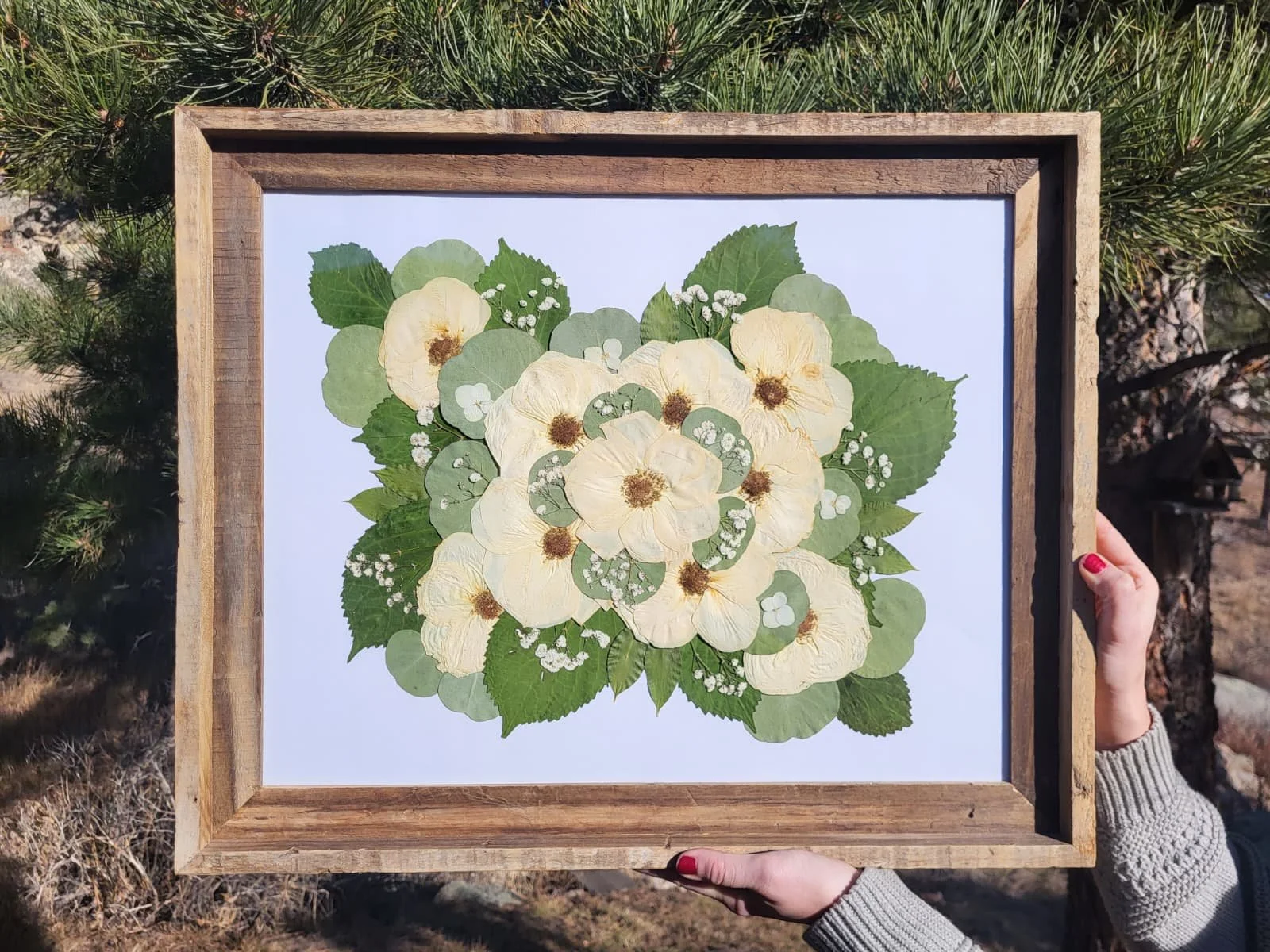 Person holding a framed botanical illustration of white flowers and green leaves outdoors.