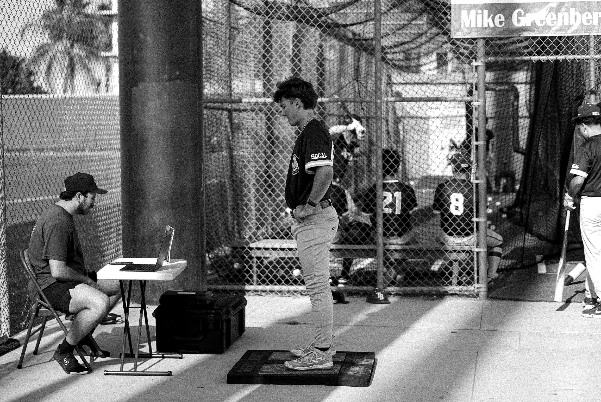 Two young boys and an adult man exercise in a gym with black walls and large mirror. The boys are jumping over orange agility rings on the black floor, while the man stands beside them, smiling. The gym has workout equipment and mirrors, and the walls are decorated with black and white graffiti-style artwork.