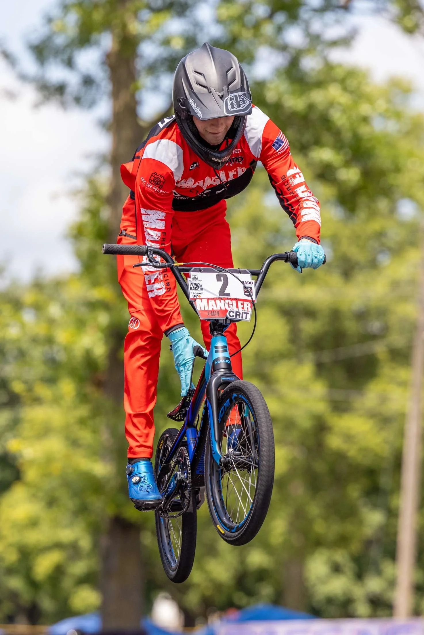 A mountain biker wearing a red and black racing suit and helmet, riding a blue bicycle in mid-air during a race in a wooded area.