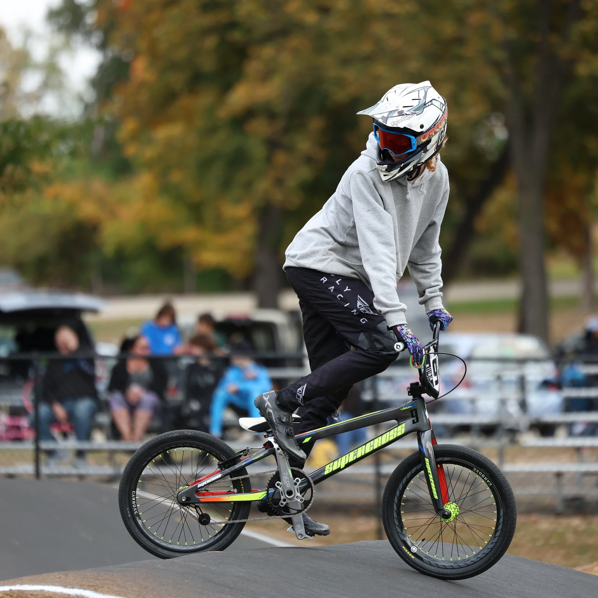 A person wearing a helmet, goggles, a gray hoodie, and black pants riding a BMX bike on a dirt track, with spectators in the background.