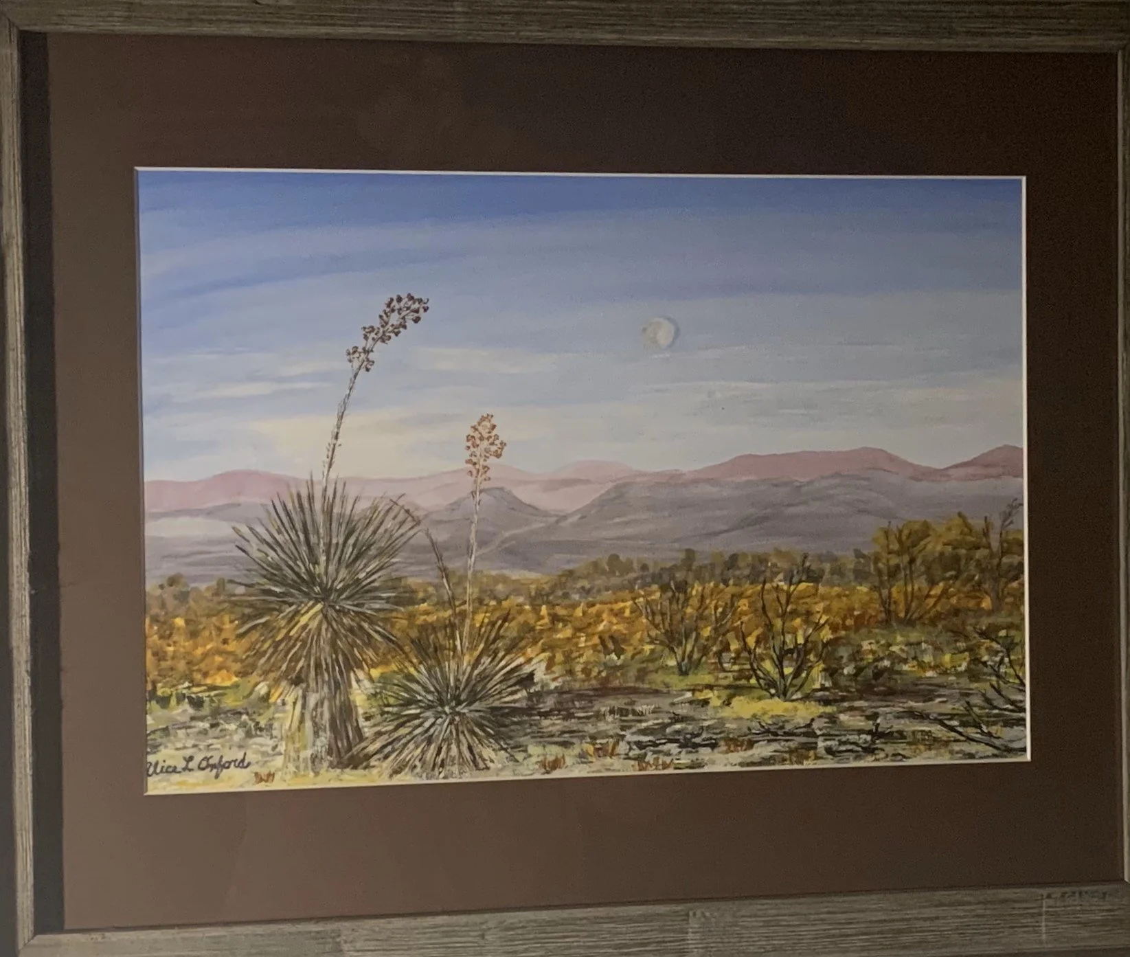 Alice Oxford's process photo of landscape painting depicting a desert scene with yucca plants in the foreground, a rugged terrain with yellow and green vegetation, distant purple mountains, and a blue sky with a faint moon or sun.